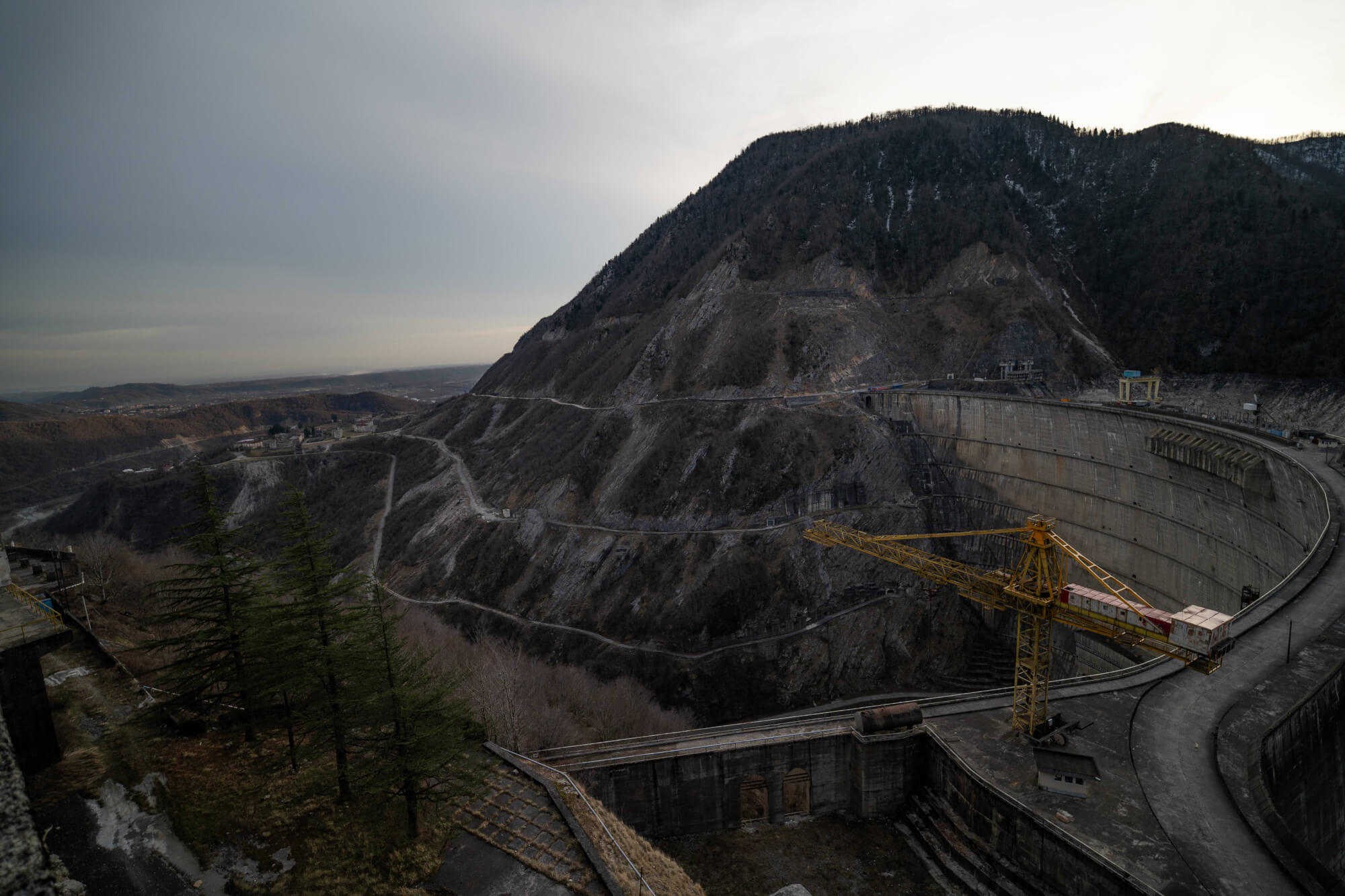 A view of a large dam set among steep, barren mountains under a cloudy sky. In the foreground, sparse trees and remnants of structures are visible. A yellow crane is positioned near the dam, suggesting signs of previous activity in the area.