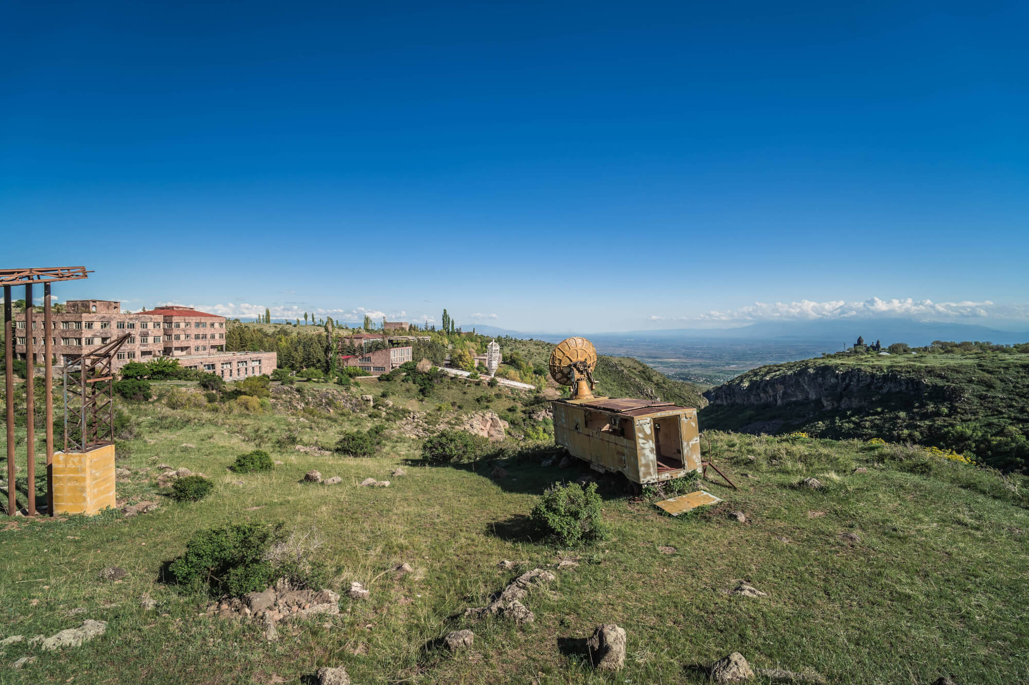 A hillside with a dilapidated structure and a satellite dish in the foreground, surrounded by lush green scenery and old buildings partially hidden by trees, under a clear blue sky.