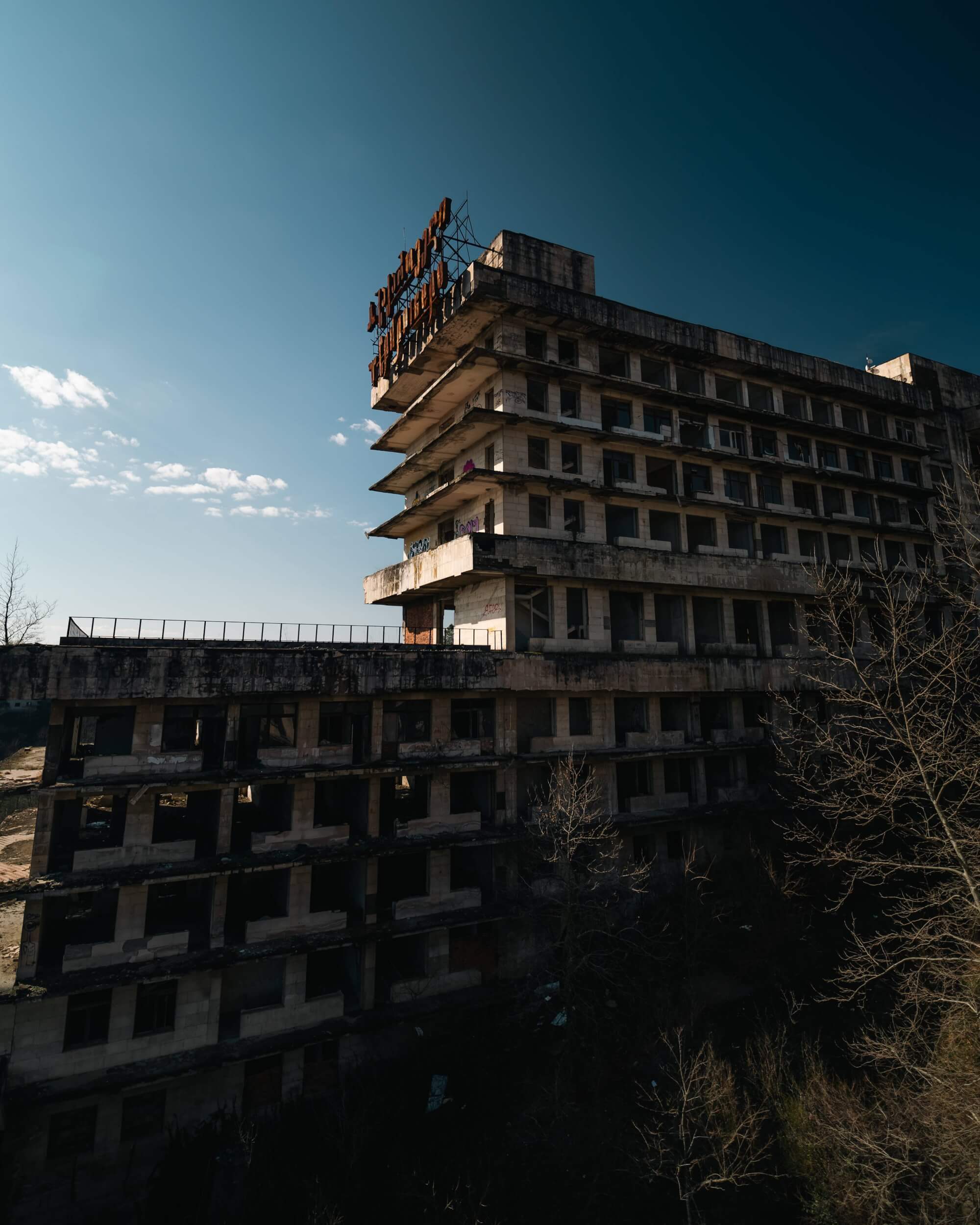 A rundown hotel building with a rusty sign on the roof, set against a clear blue sky. The structure has many empty windows and is partially surrounded by bare trees.
