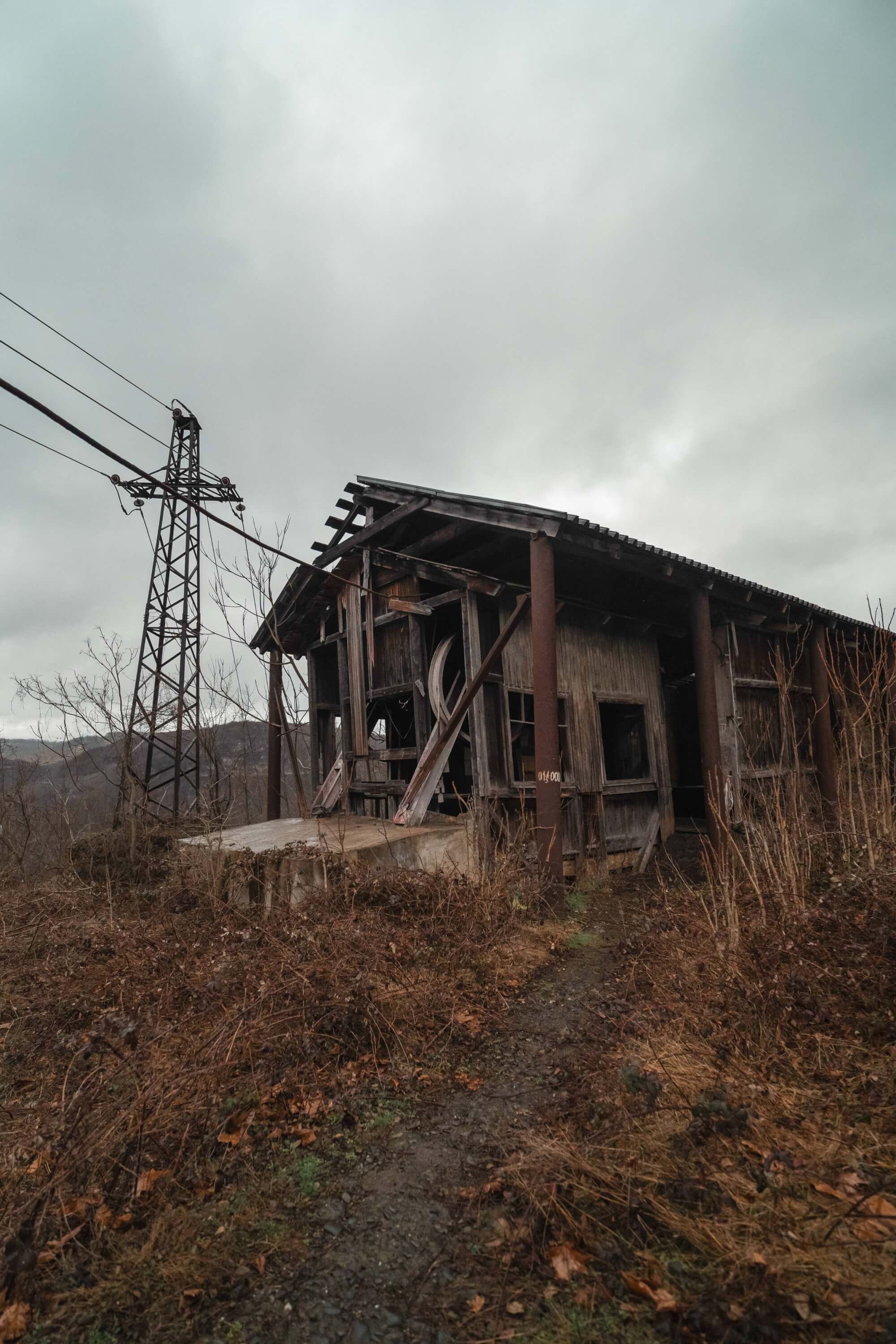 A run-down wooden structure with broken windows and a collapsing roof, surrounded by dry shrubs and a muddy path. A power line stands nearby under a cloudy sky.