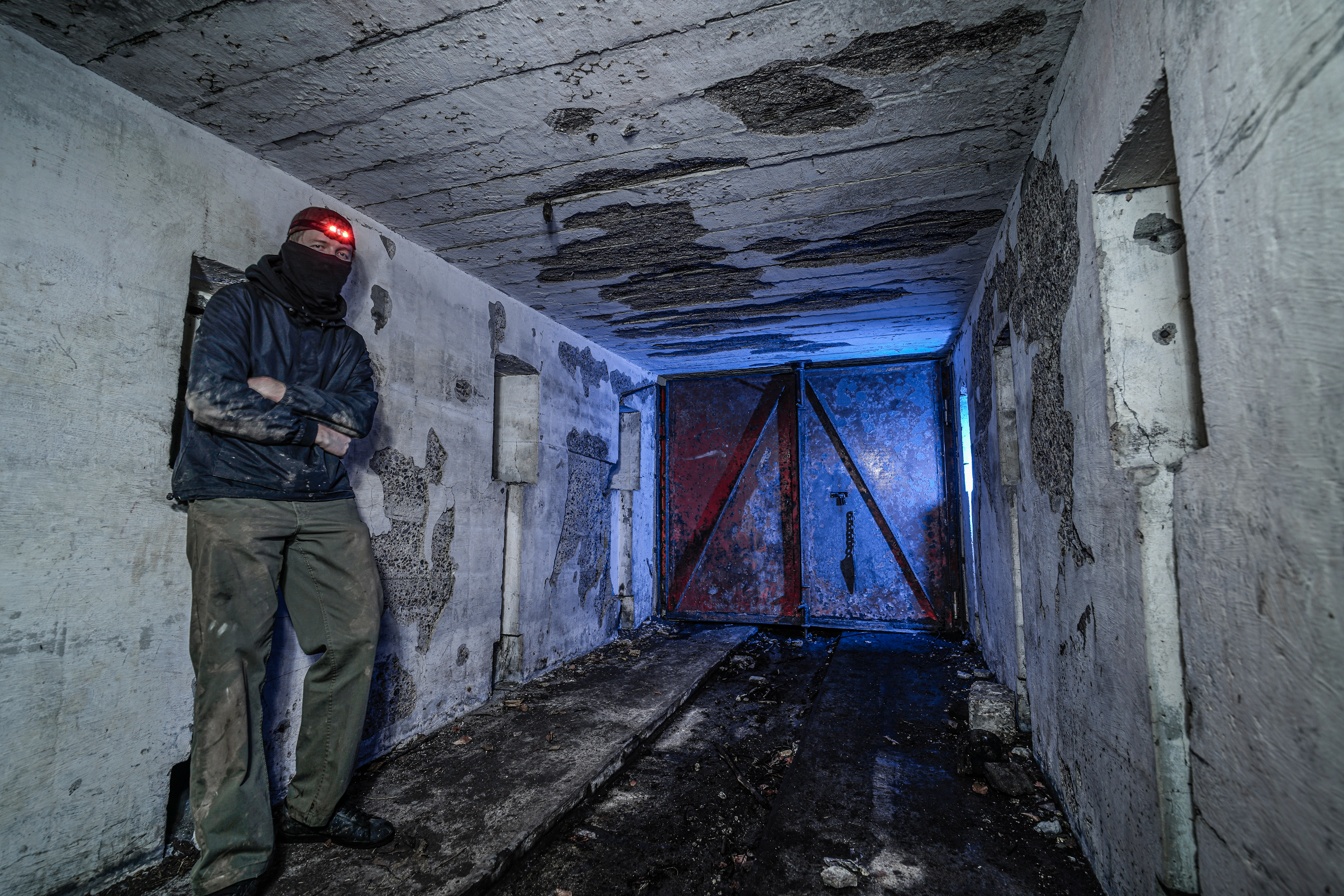 A person in a dark hoodie and mask stands against a wall in a neglected concrete corridor. A dim red light from their headlamp illuminates part of their face. The corridor features a locked red door in the background, with peeling walls and a dirty floor.