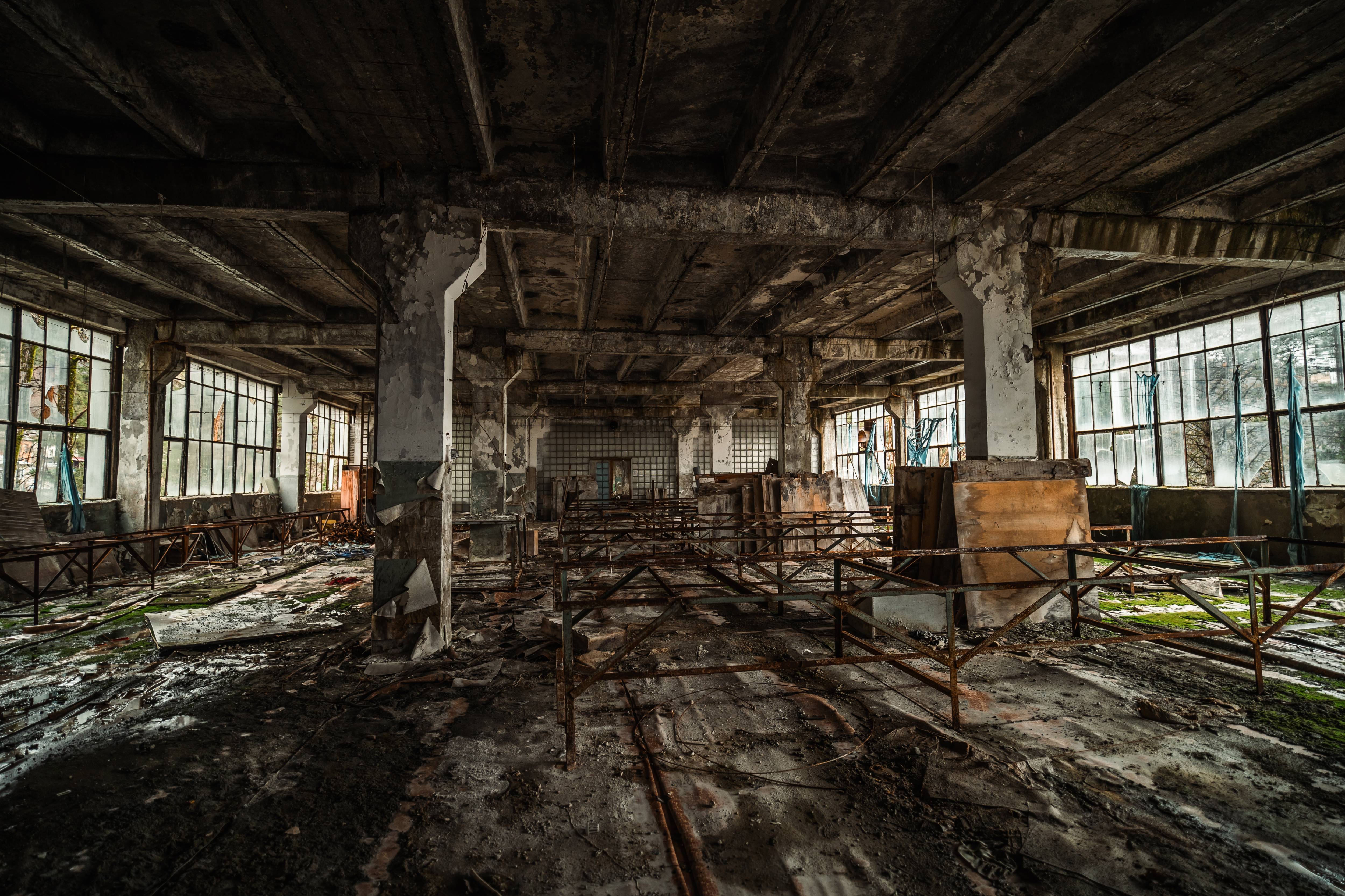 An abandoned factory interior with cracked concrete floors and tall windows. Rusty metal frames are scattered throughout the space, with patches of green moss visible. The atmosphere is one of decay, with shadows cast by the broken windows.