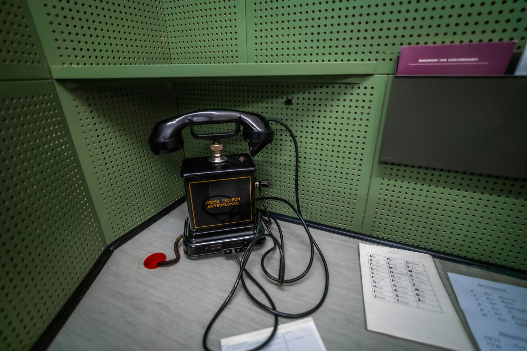 A vintage black telephone with tangled wires sits in the corner of a room with green perforated walls. Nearby, a few sheets of paper with numbers and letters are visible on a light gray surface.
