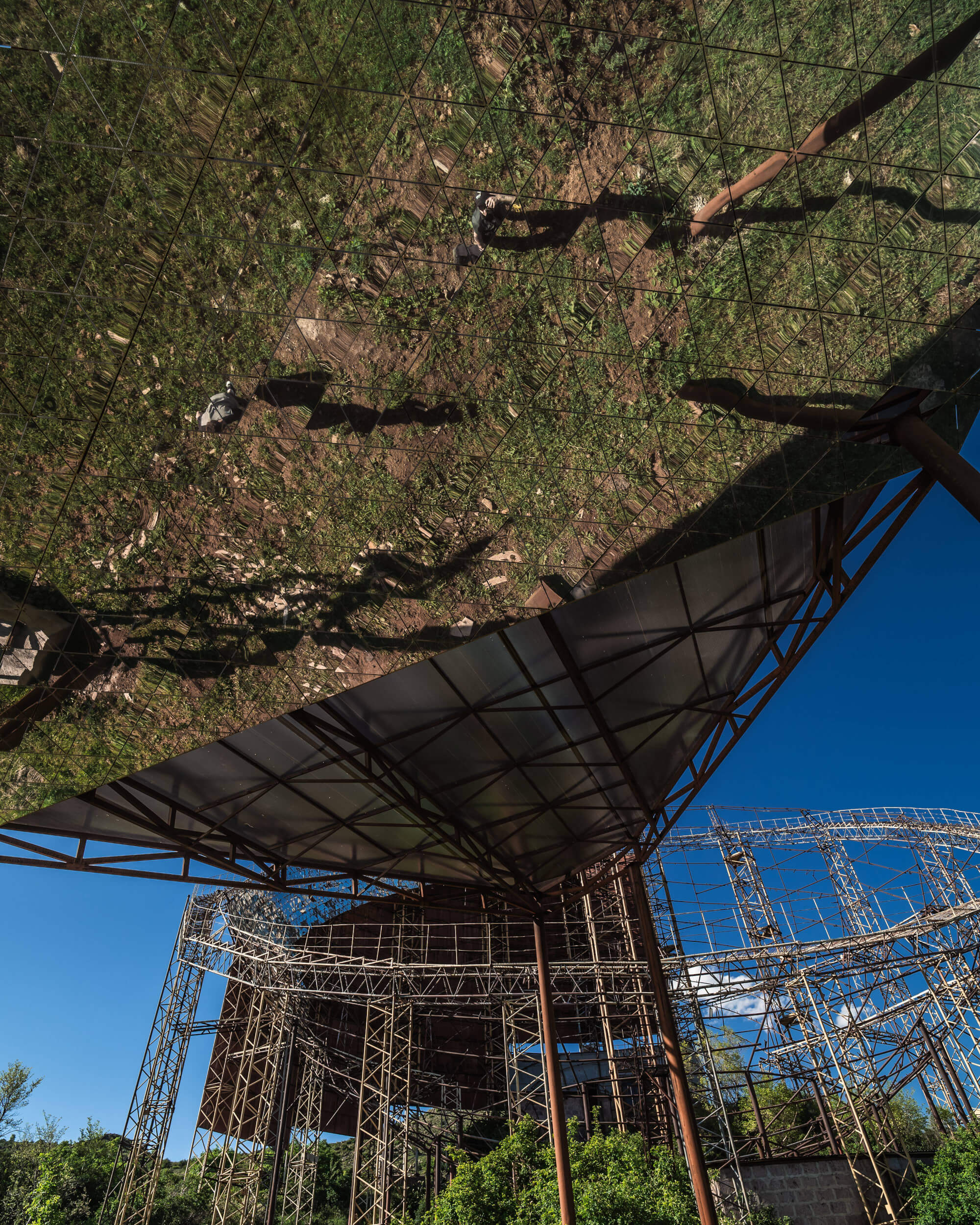 A reflective surface in the foreground shows greenery and soil, while the background features an incomplete metallic structure and a blue sky.