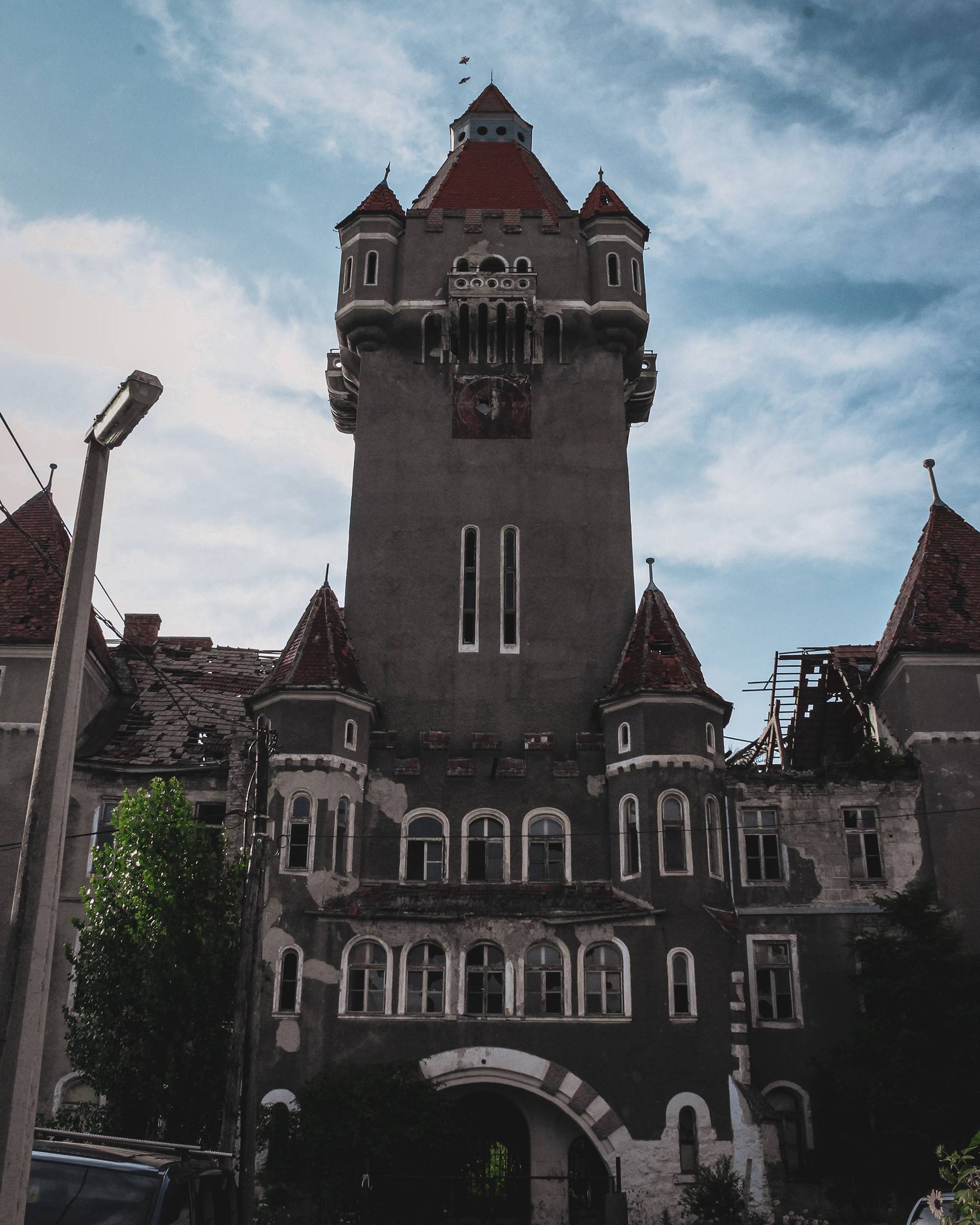 A tall, abandoned building with a gray exterior and red roof accents, featuring intricate windows and a partially collapsed rooftop, surrounded by greenery and a cloudy sky.