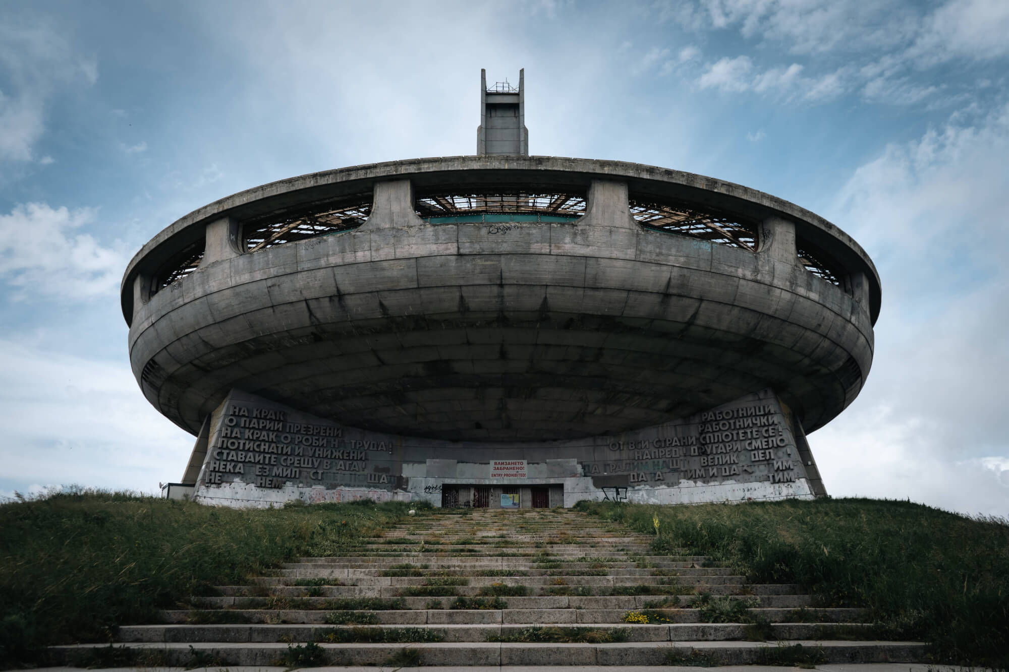 Low-angle view of an abandoned circular concrete monument with large inscriptions, surrounded by grass and a cloudy sky.