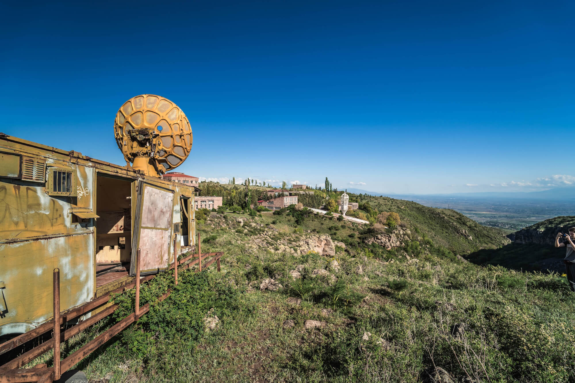 An abandoned vehicle with a large yellow satellite dish on top, surrounded by green hills and distant buildings under a clear blue sky.