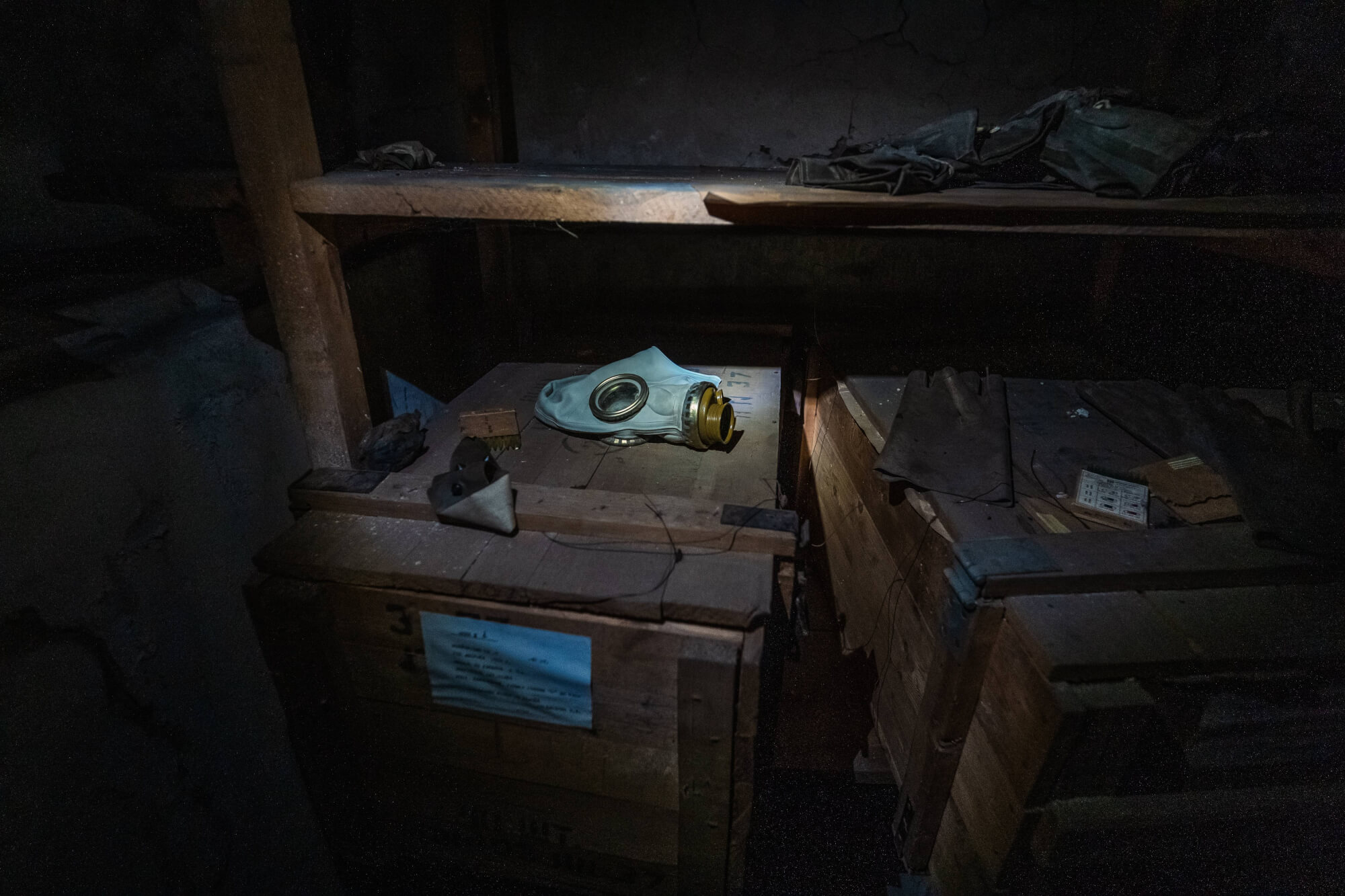 A gas mask placed on a wooden crate in a dark storage area with scattered gloves and pieces of fabric around it, illuminated by a narrow beam of light.