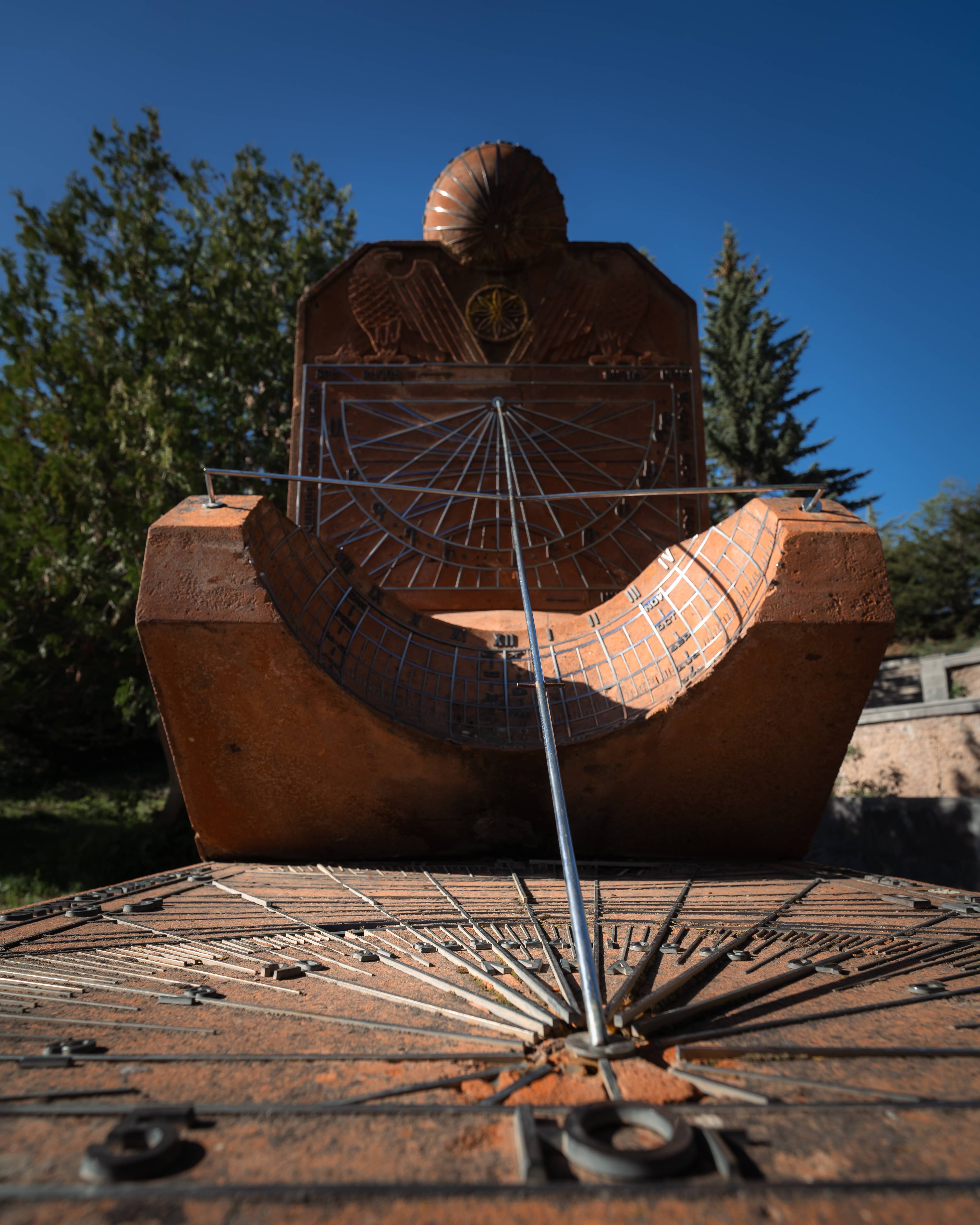 Close-up of a sundial made of metal and stone, with a shadow-casting gnomon and various engraved markings, set against a clear blue sky and blurred greenery in the background.