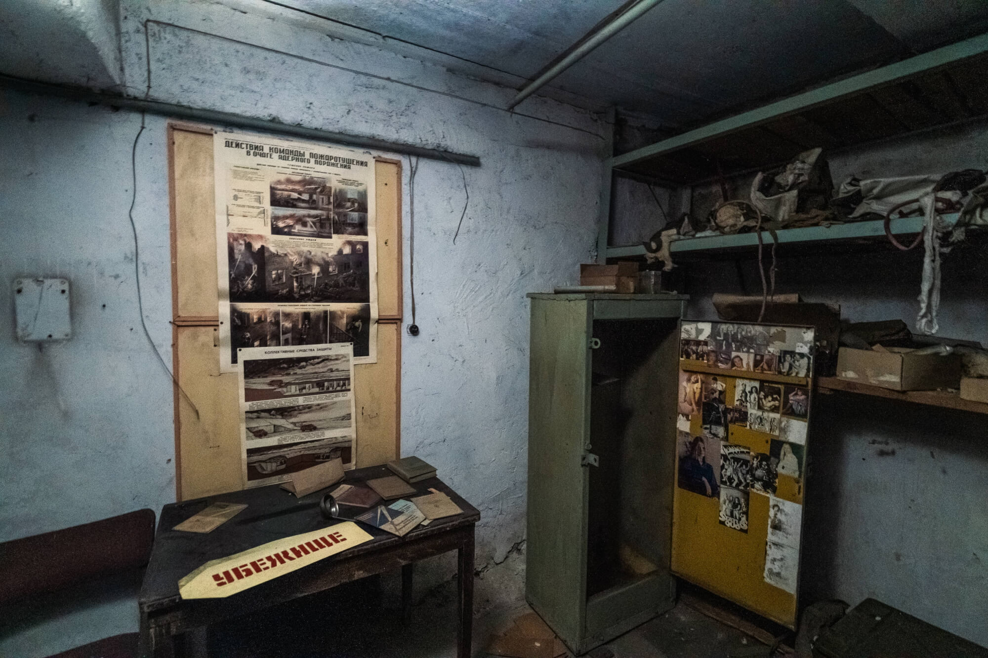 An old, dim room with a wooden table covered in scattered papers and a cylindrical object. A vintage emergency response poster is pinned to the wall, and a yellow cabinet with photographs is visible on the right, accompanied by shelves holding various items.