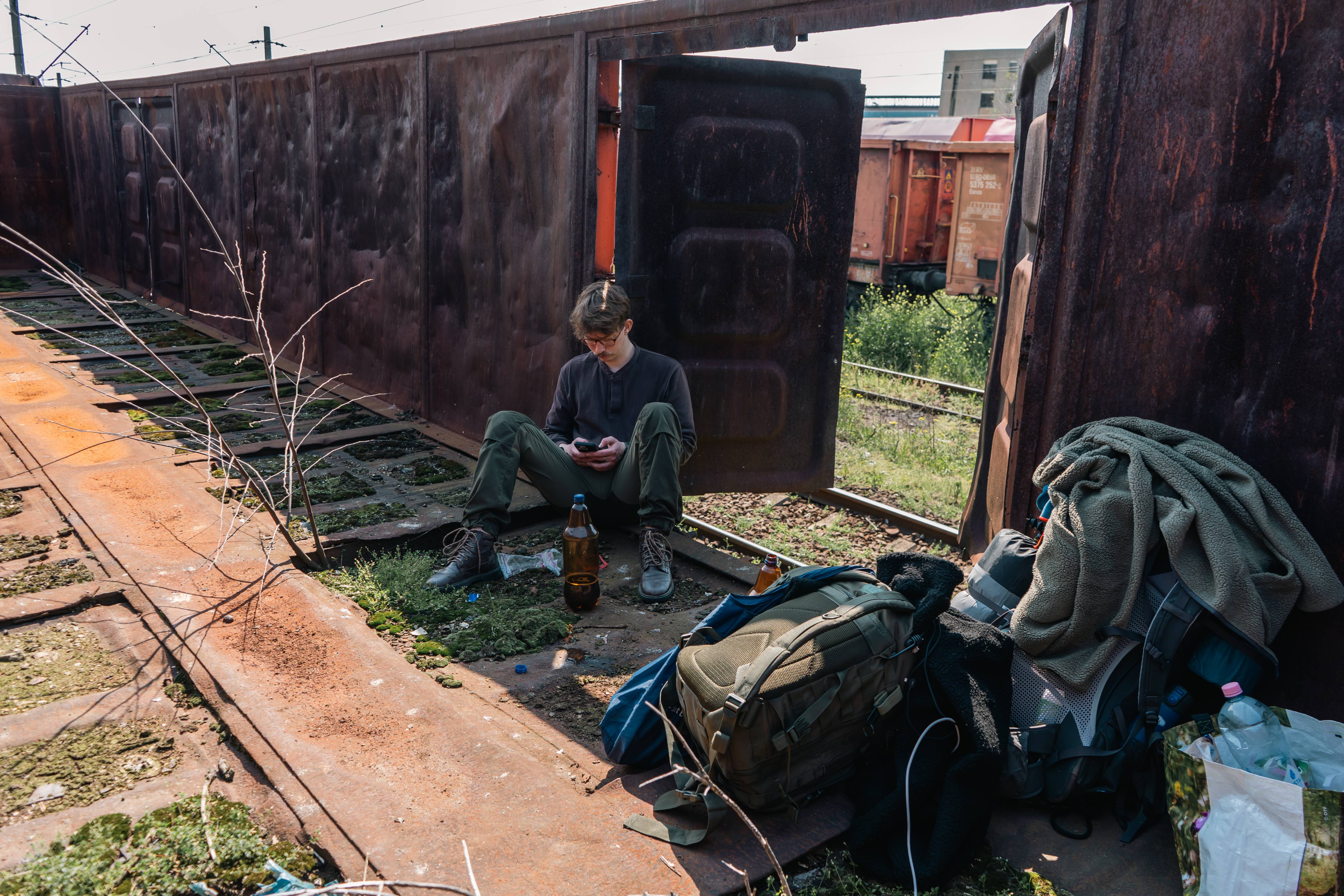 A young person sits on the ground in an abandoned train yard, looking at their phone, surrounded by bags and empty bottles.