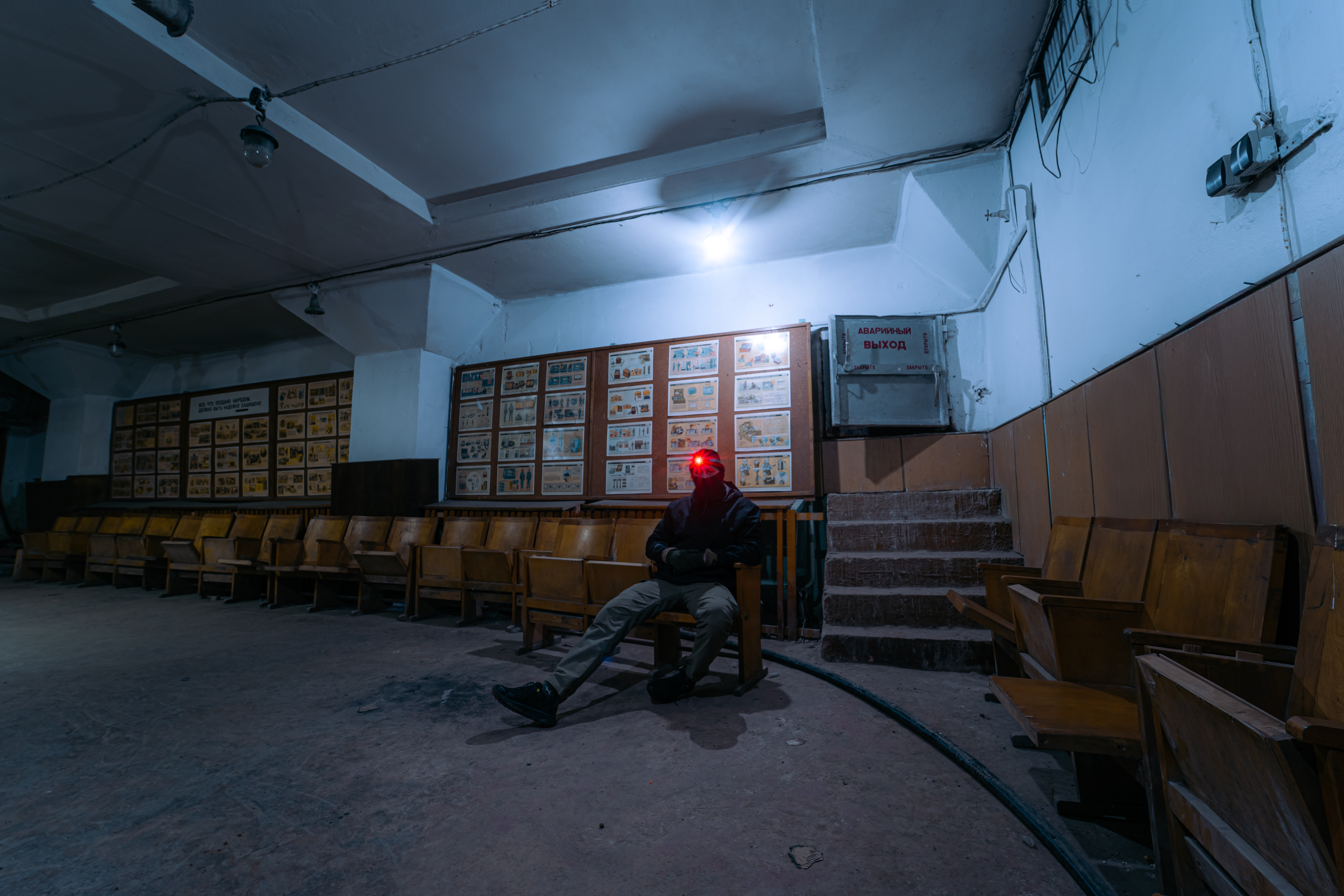A dimly lit abandoned waiting area with wooden benches. A person wearing dark clothing and a red headlamp sits alone. The walls feature faded instructional posters, and a few steps lead further into the space.
