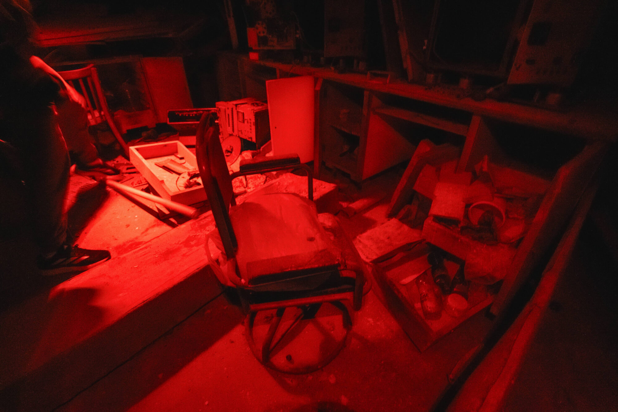 A dimly lit room with red lighting featuring a worn-out chair surrounded by debris, including broken boxes and discarded items, creating an eerie and abandoned atmosphere.
