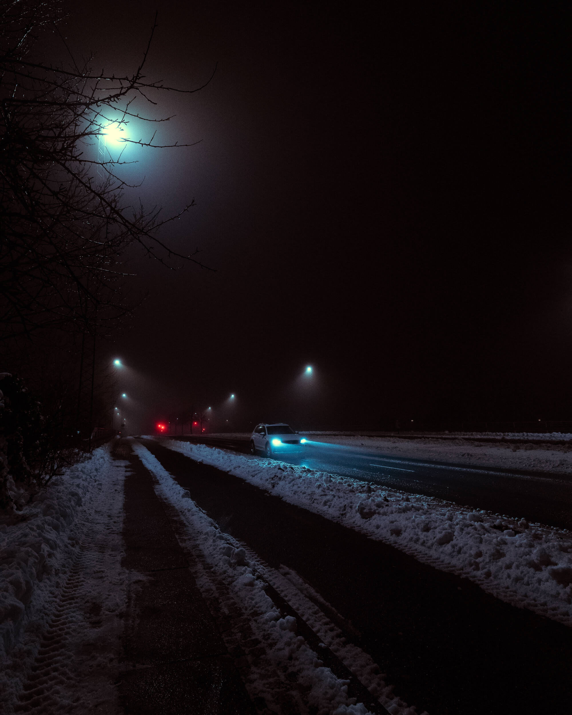 A foggy nighttime street scene with snow on the ground. A car with bright headlights drives along a dark road, flanked by streetlights illuminating the fog. Bare branches are visible on the left side of the image.