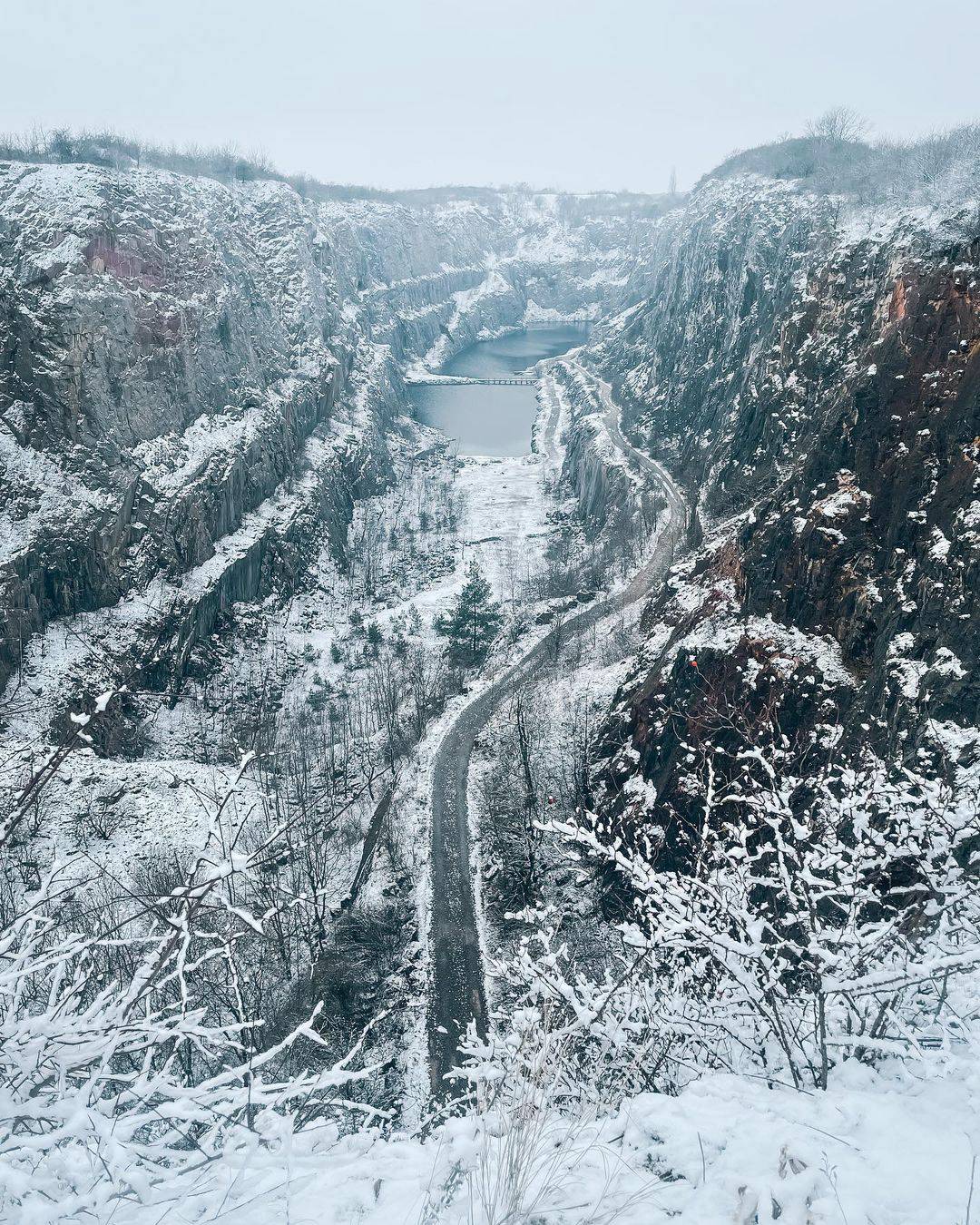 A snow-covered quarry with steep rocky cliffs, a winding path, and a frozen lake in the background under a cloudy sky.