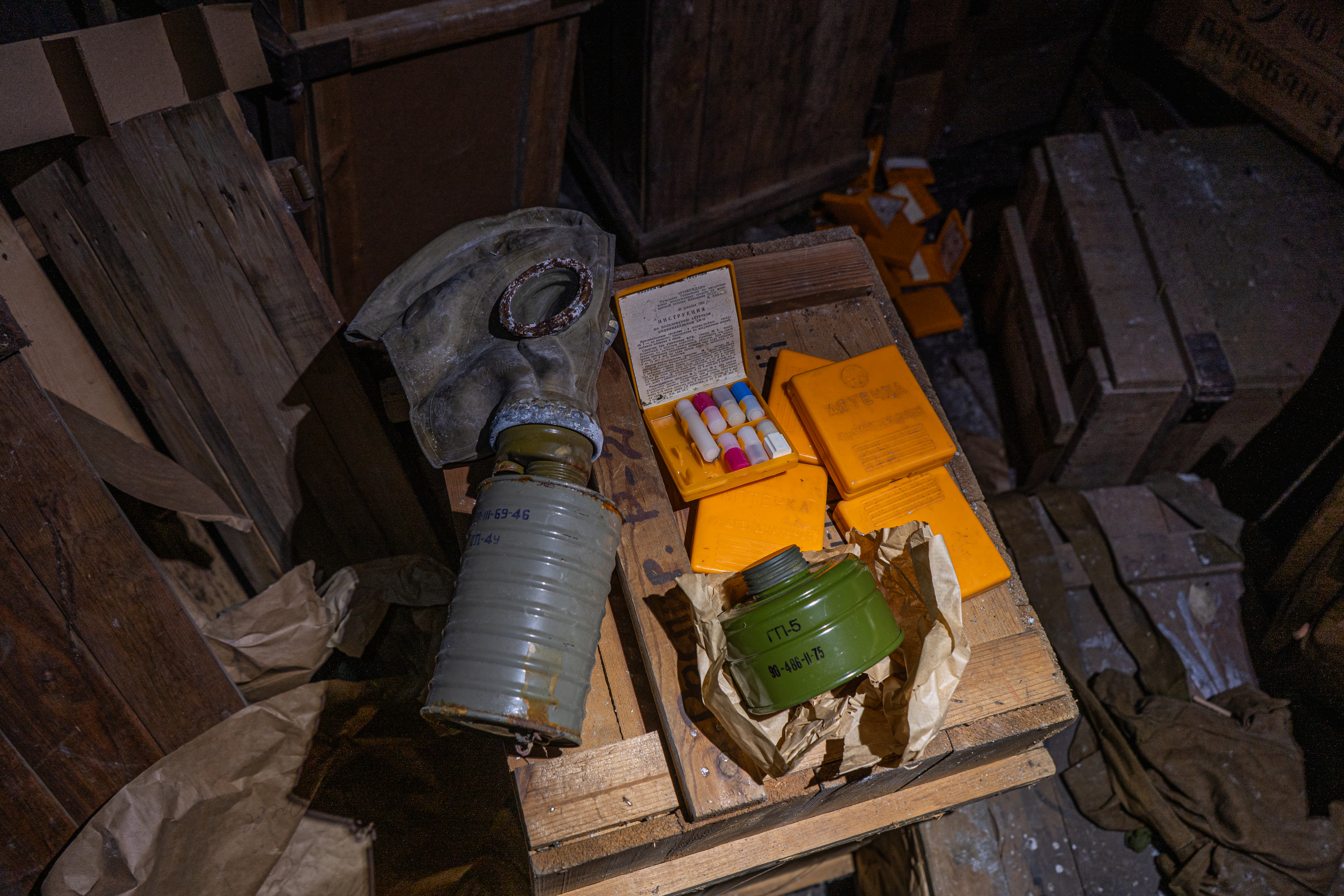 A gas mask and several yellow containers are placed on a wooden table amid a cluttered and dusty environment, with wooden crates and papers in the background.