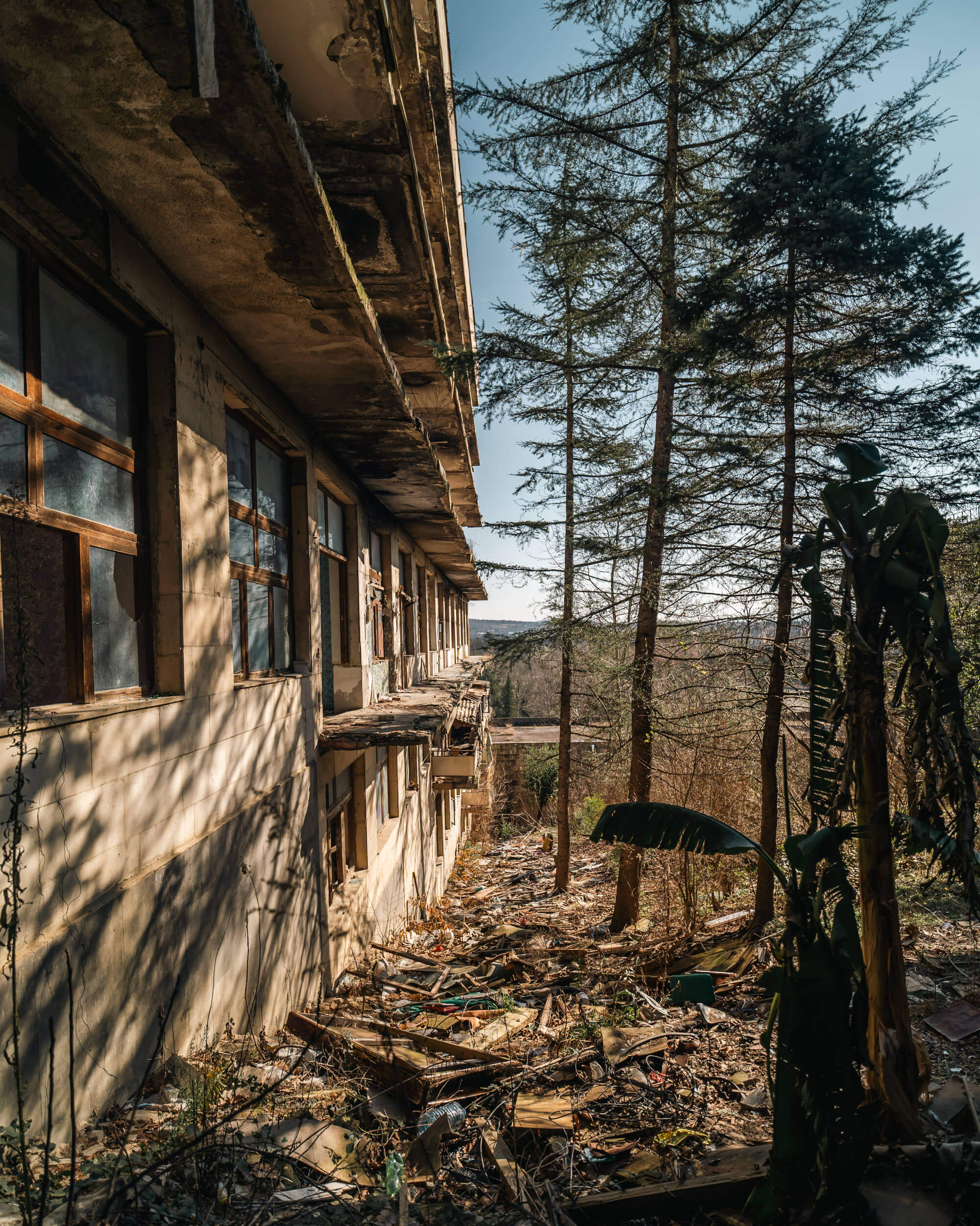 An abandoned building with broken windows and a crumbling exterior, surrounded by trees and scattered debris. Sunlight creates shadows on the wall, showcasing the deterioration of the structure.