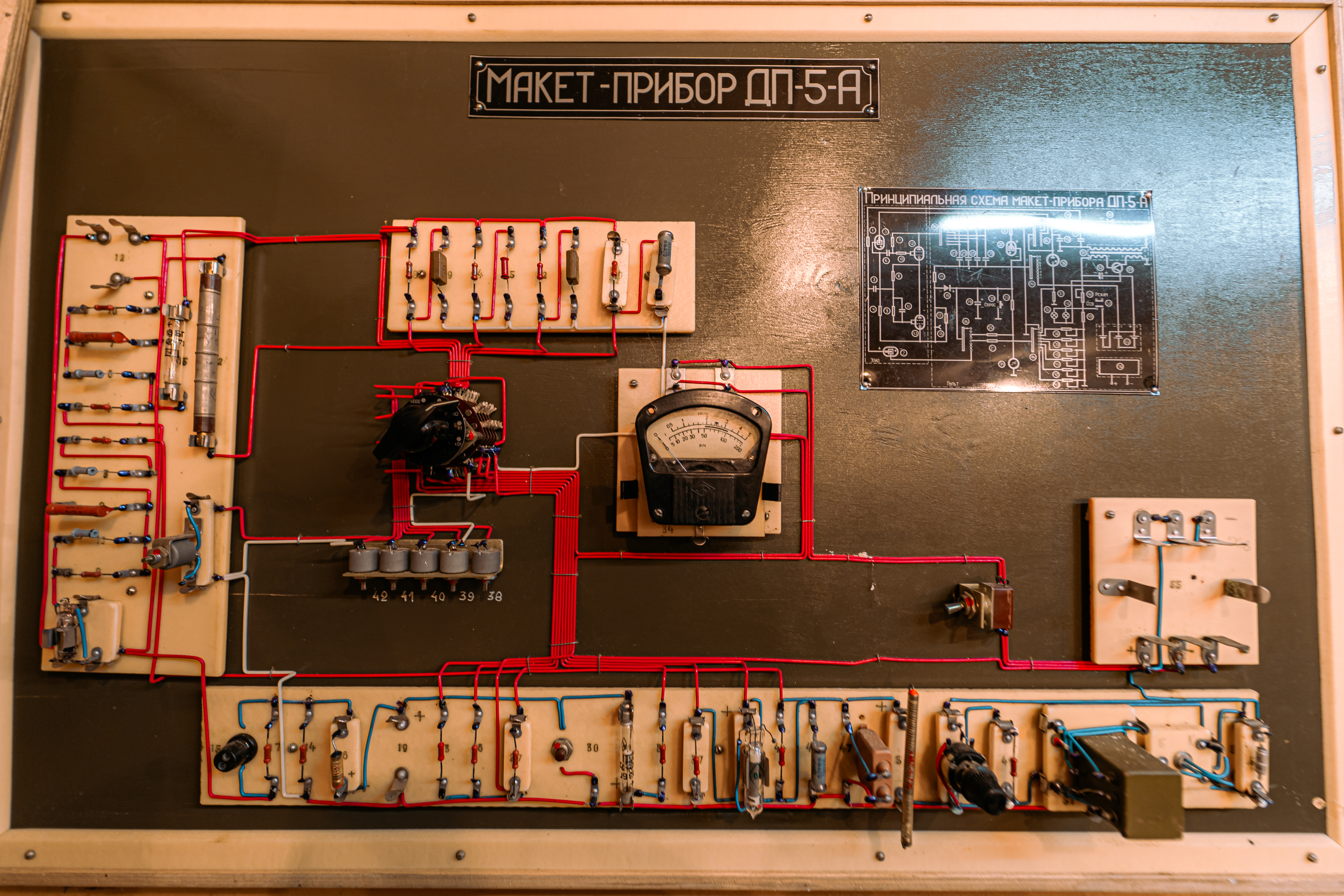 A close-up view of a model circuit board with red and blue wiring connecting various components, including a central meter and a schematic diagram in the upper right corner.