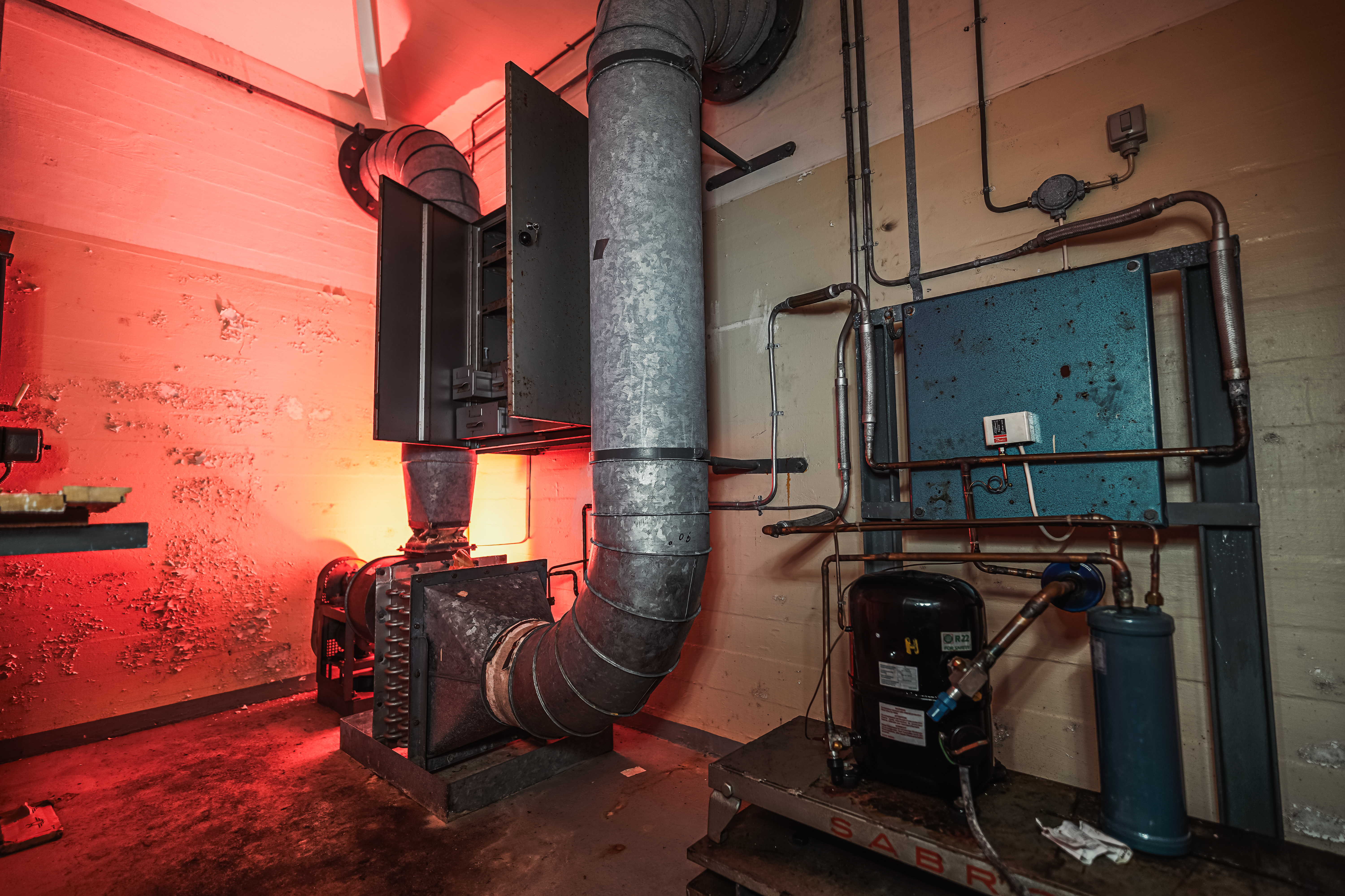 An industrial room featuring exposed metal pipes and machinery, with red light illuminating the area, creating a contrast between the cold metal and warm light.