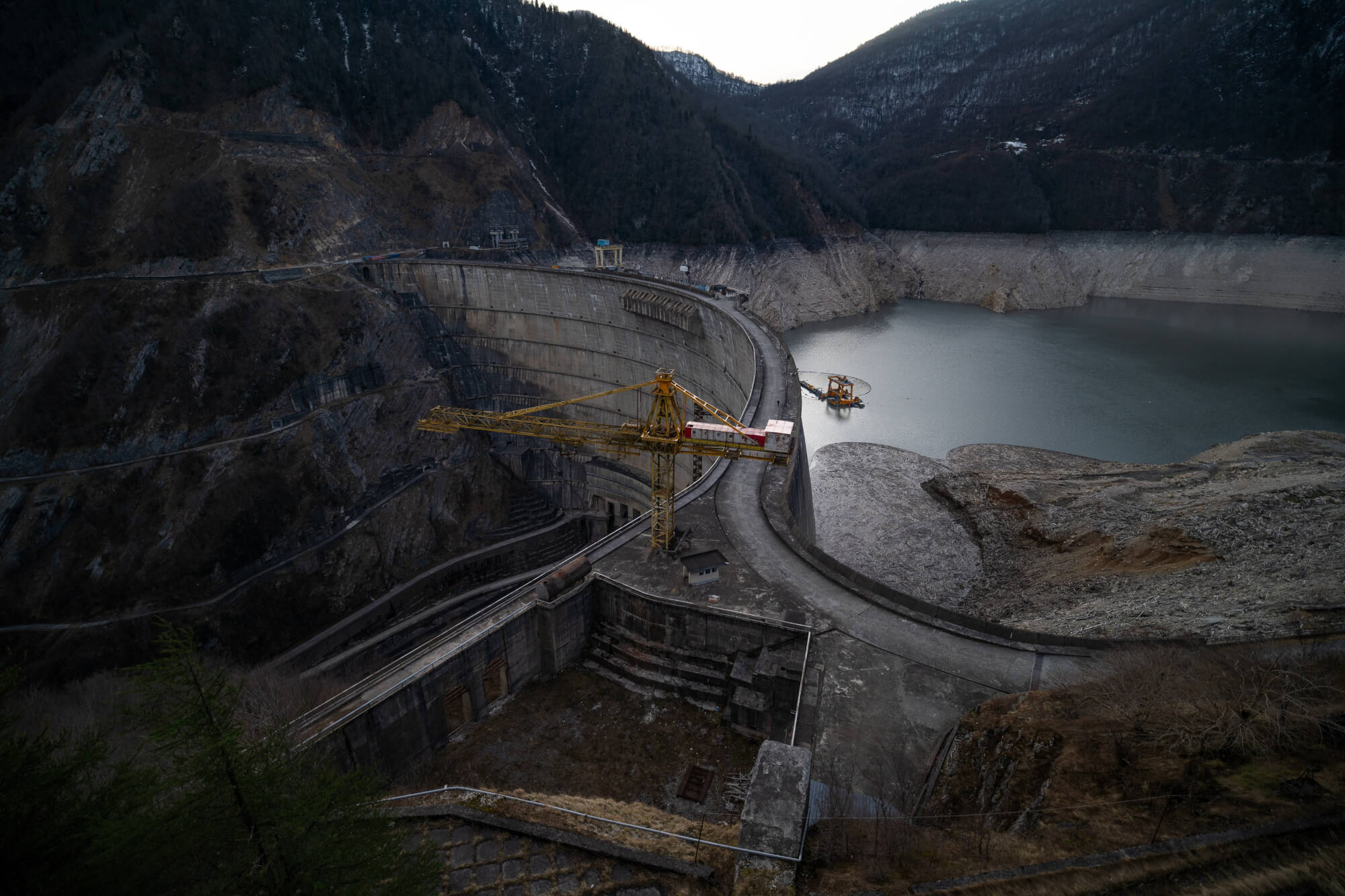 A large dam with a yellow crane on top, surrounded by mountains and a calm body of water. The landscape is mostly barren and reflects a cloudy sky.