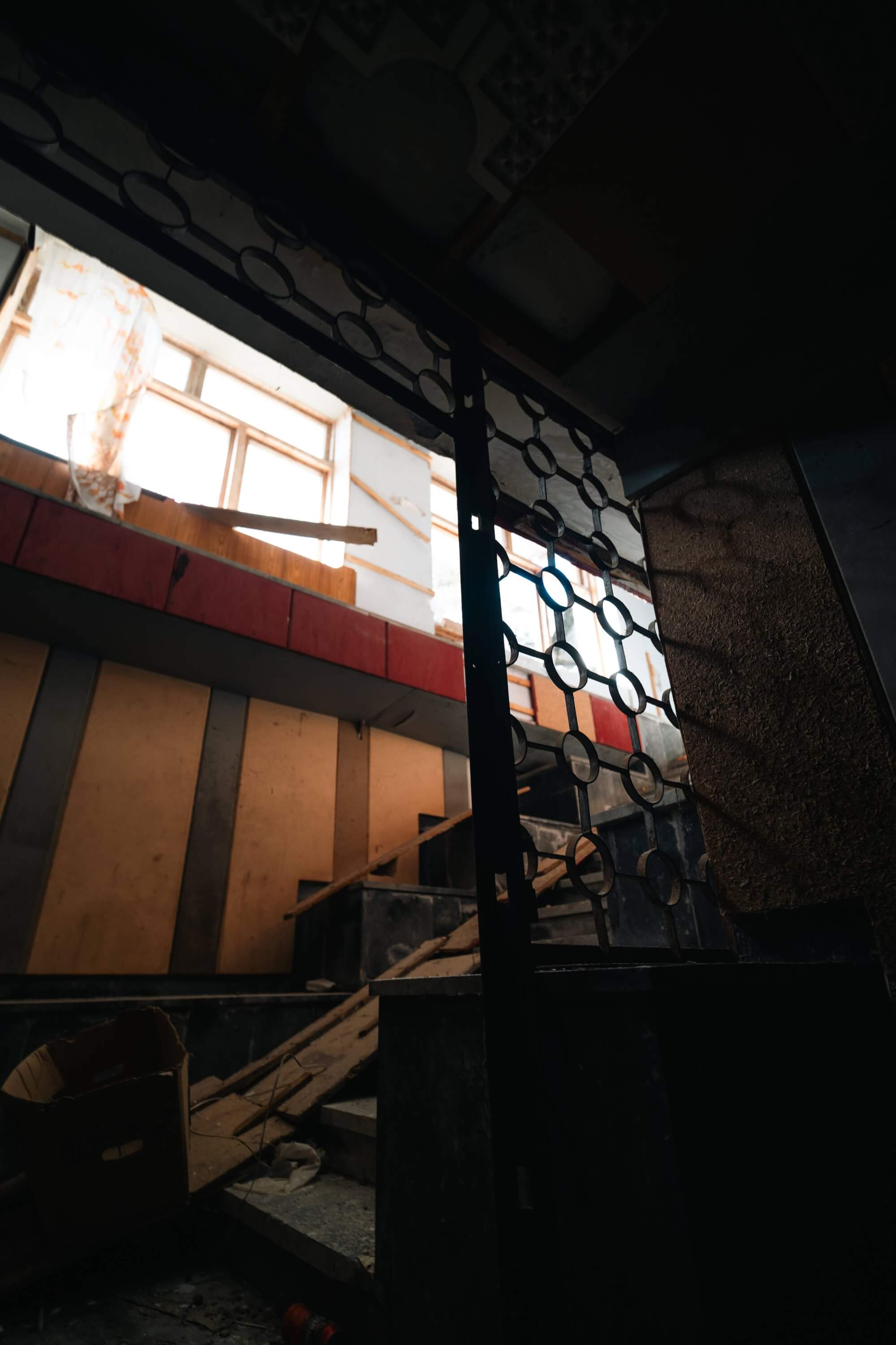 A dark staircase with a wrought iron gate in the foreground, leading to an upper level bathed in soft light from a window. The scene includes wooden panels, concrete walls, and scattered debris, conveying a sense of abandonment.