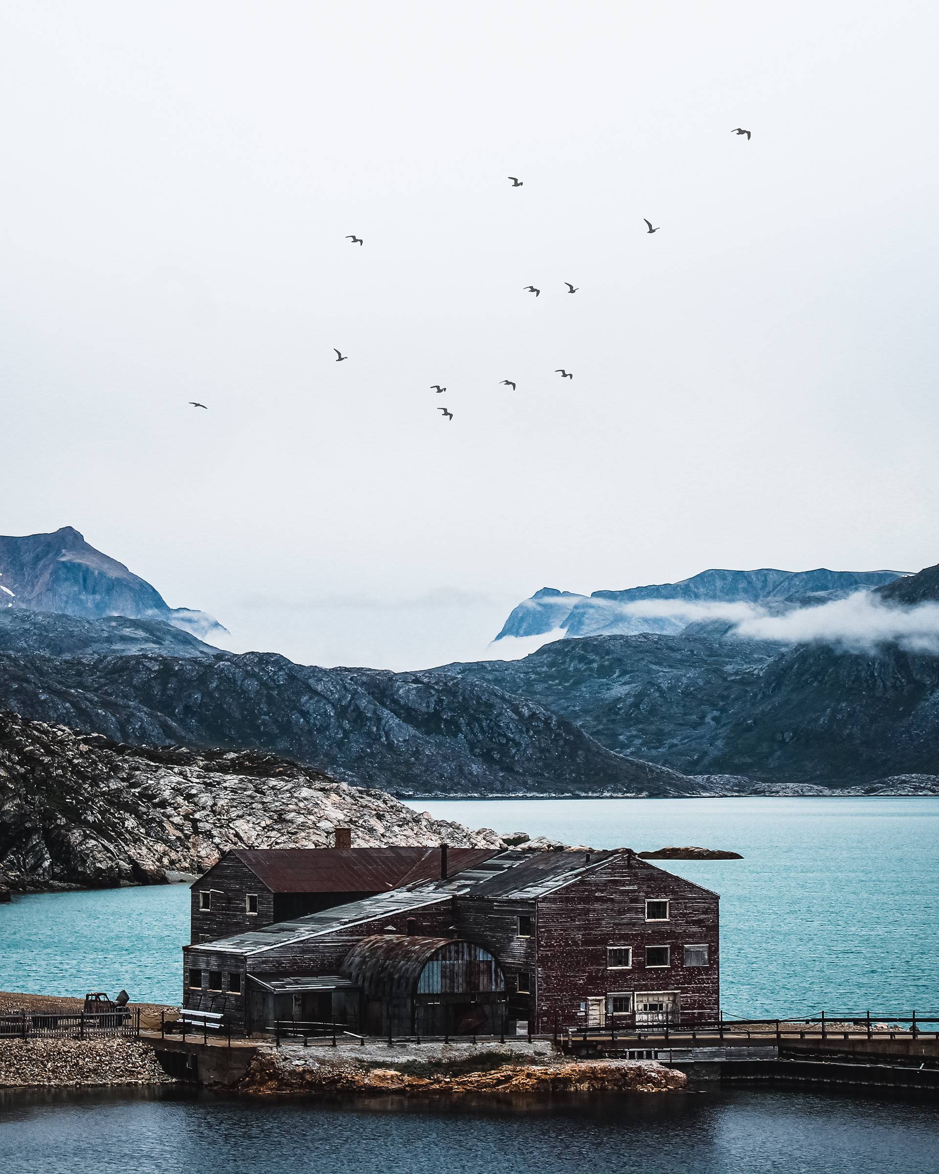 An old wooden building by a calm turquoise bay, with rugged, misty mountains in the background and a flock of birds in the sky.