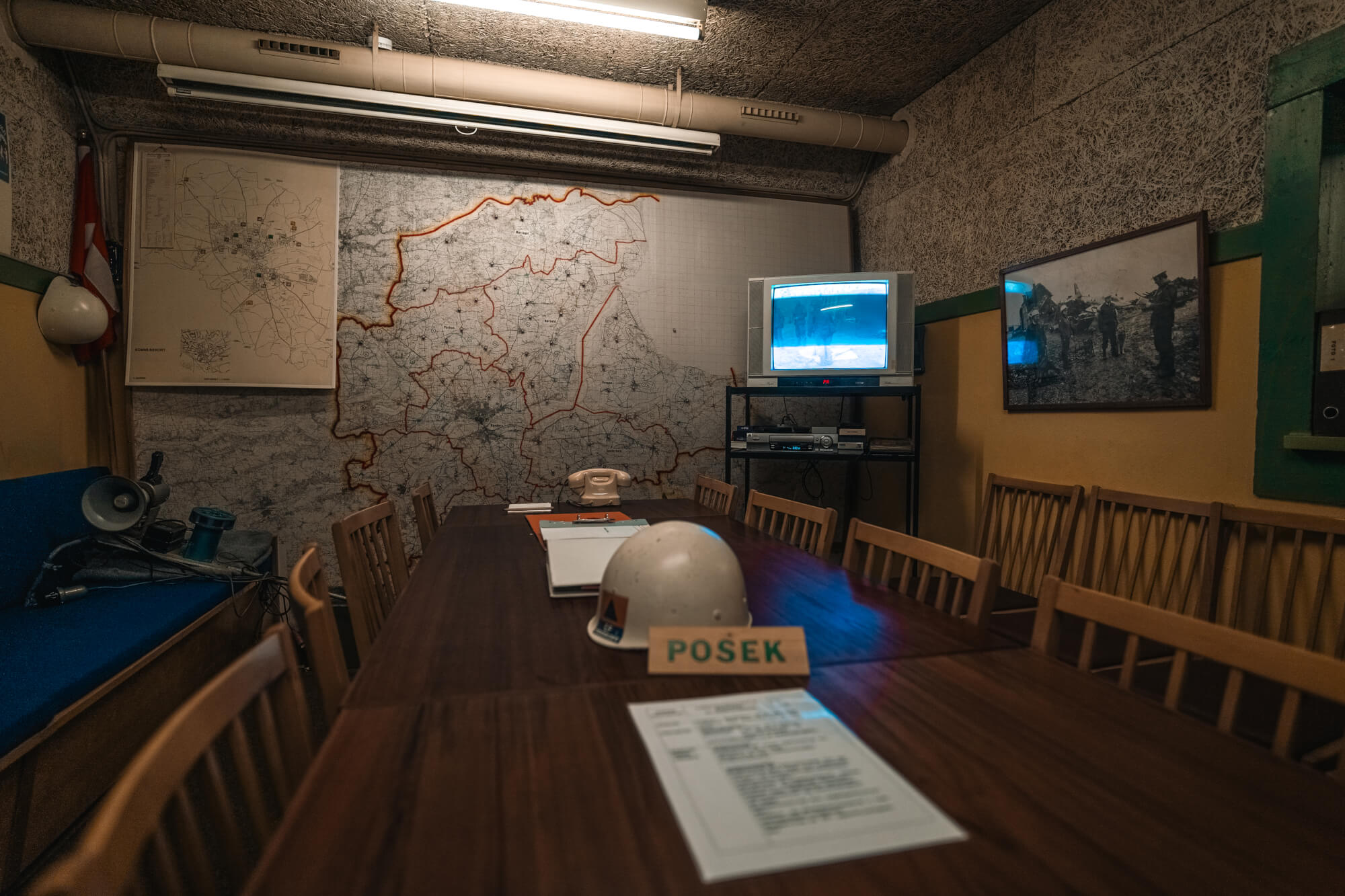 A command room featuring a long wooden table, documents, and a hard hat with the name 'POŠEK.' The back wall has a large map with orange outlines, while a vintage television displays static. Framed photographs are on the sidewall, contributing to the room's historical ambiance.