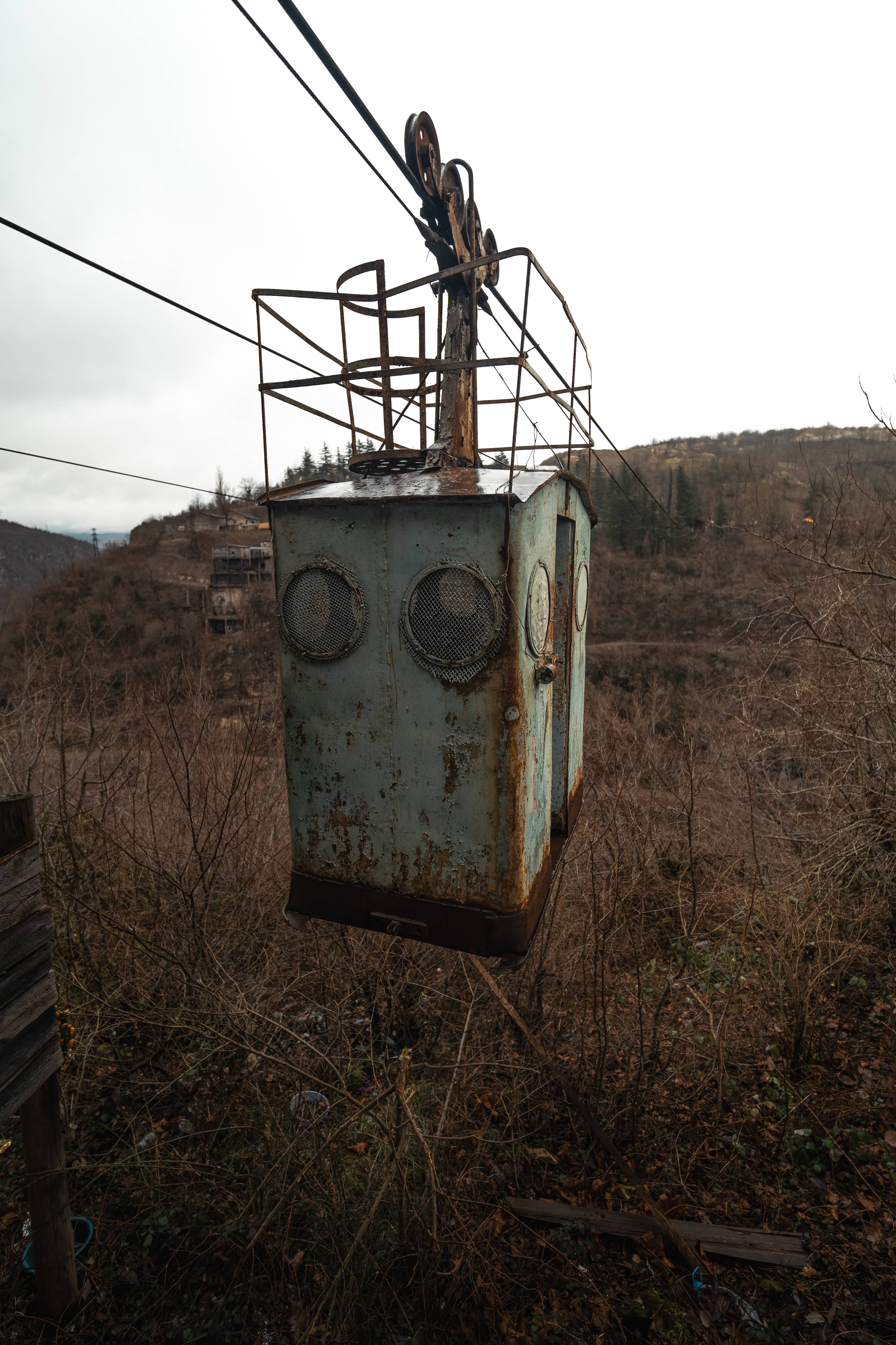 An old, rusted cable car hanging from wires above a barren landscape, surrounded by dry branches and hills in the background under a cloudy sky.