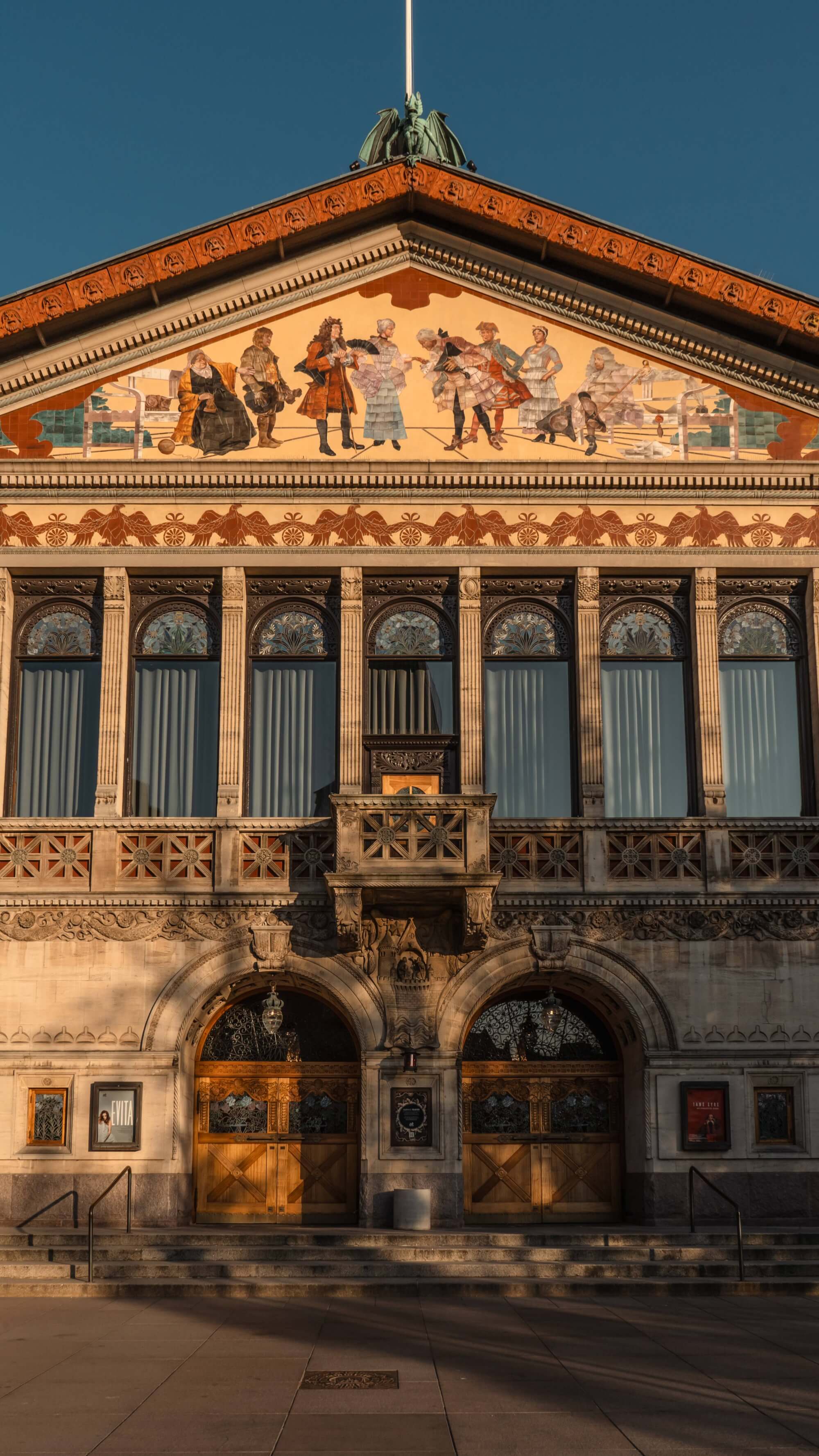 A historic building facade with ornate details. The upper portion features colorful murals of figures, while the lower part has grand arched windows and wooden doors. The scene is illuminated by warm golden light, set against a clear blue sky.