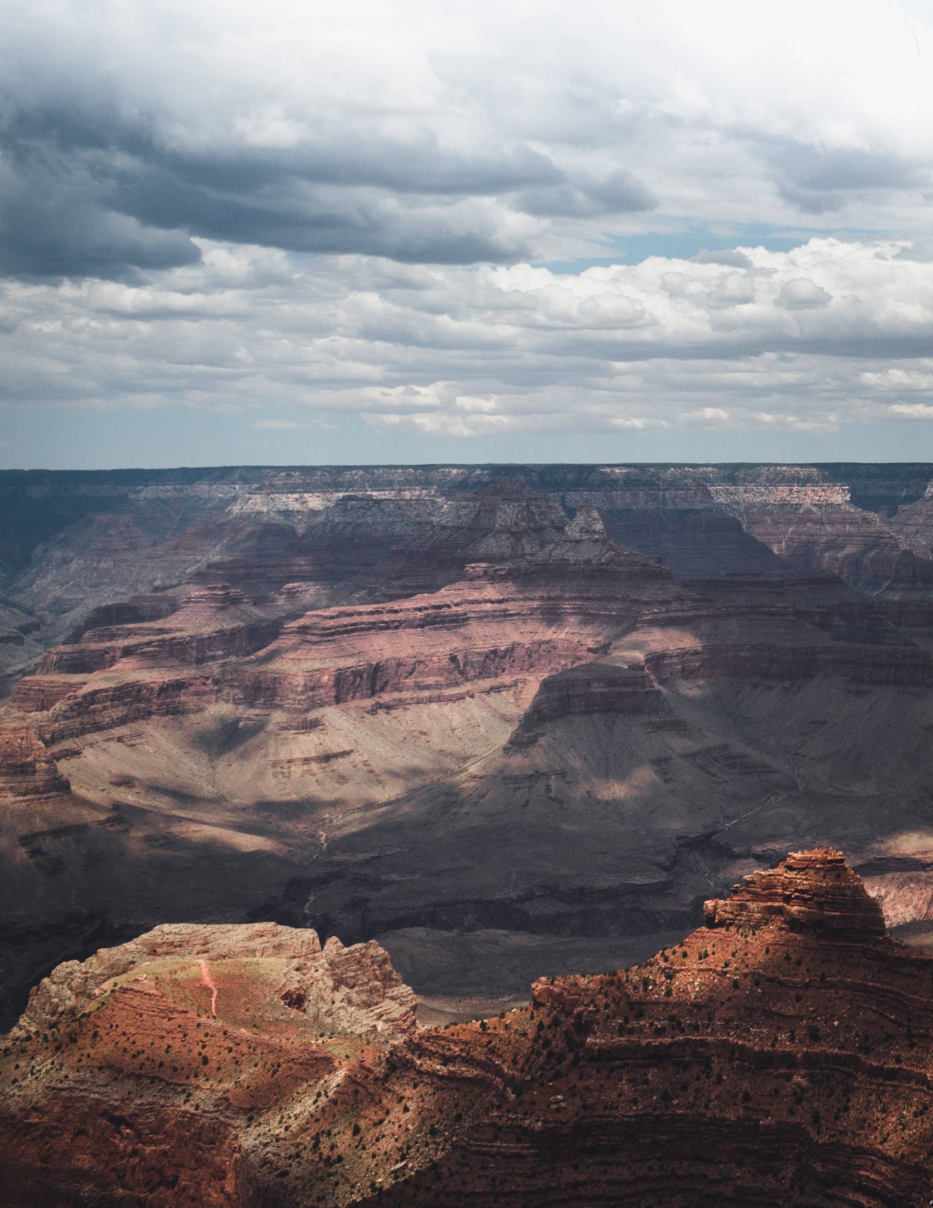 A panoramic view of the Grand Canyon featuring layered rock formations in various shades of red and brown, under a cloudy sky with patches of blue peeking through.