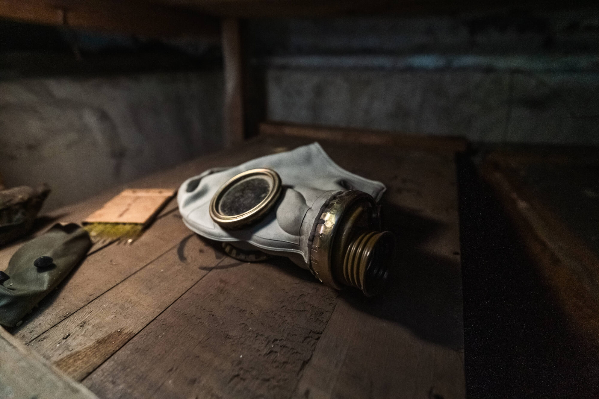A close-up of a weathered gas mask lying on a rustic wooden surface, with a dark green item and a wooden plank nearby, set in a dimly lit environment.