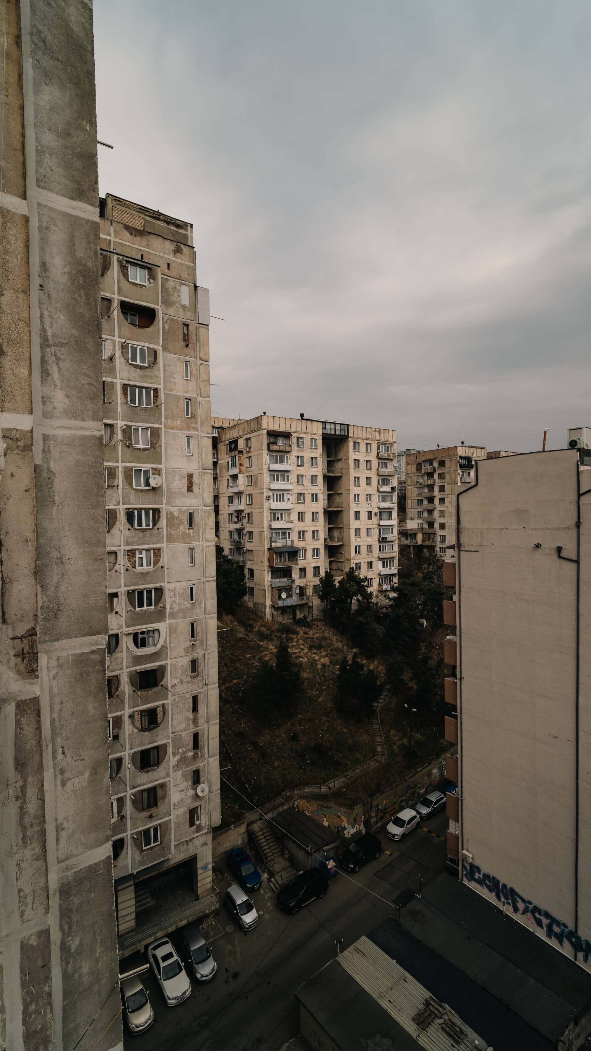 A view of concrete apartment buildings in a gray urban setting, with a hillside and trees in the background and parked cars on the street.