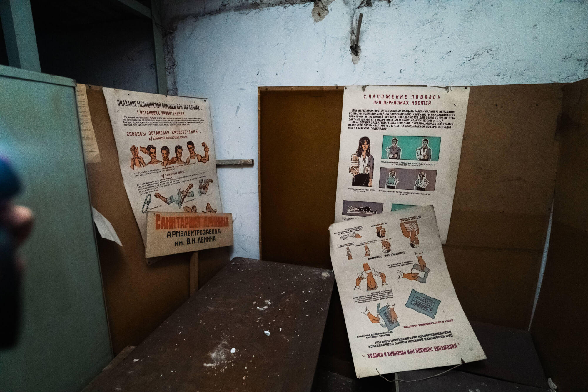 An interior view of a neglected room featuring old instructional posters on a wall. The posters contain illustrations and text related to health topics. A dusty wooden table is in the foreground.
