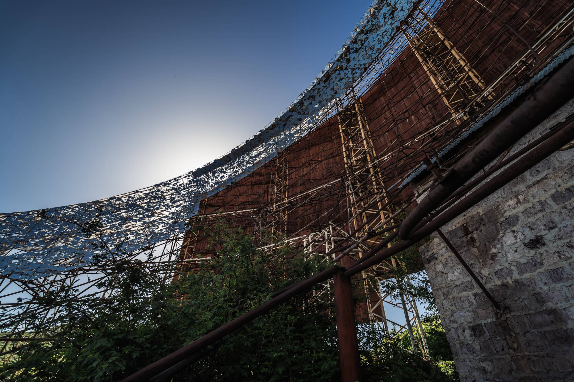A close-up view from below an abandoned curved structure with exposed scaffolding, illuminated by sunlight from behind. Overgrown vegetation is visible at the base, contrasted against the backdrop of a clear blue sky.