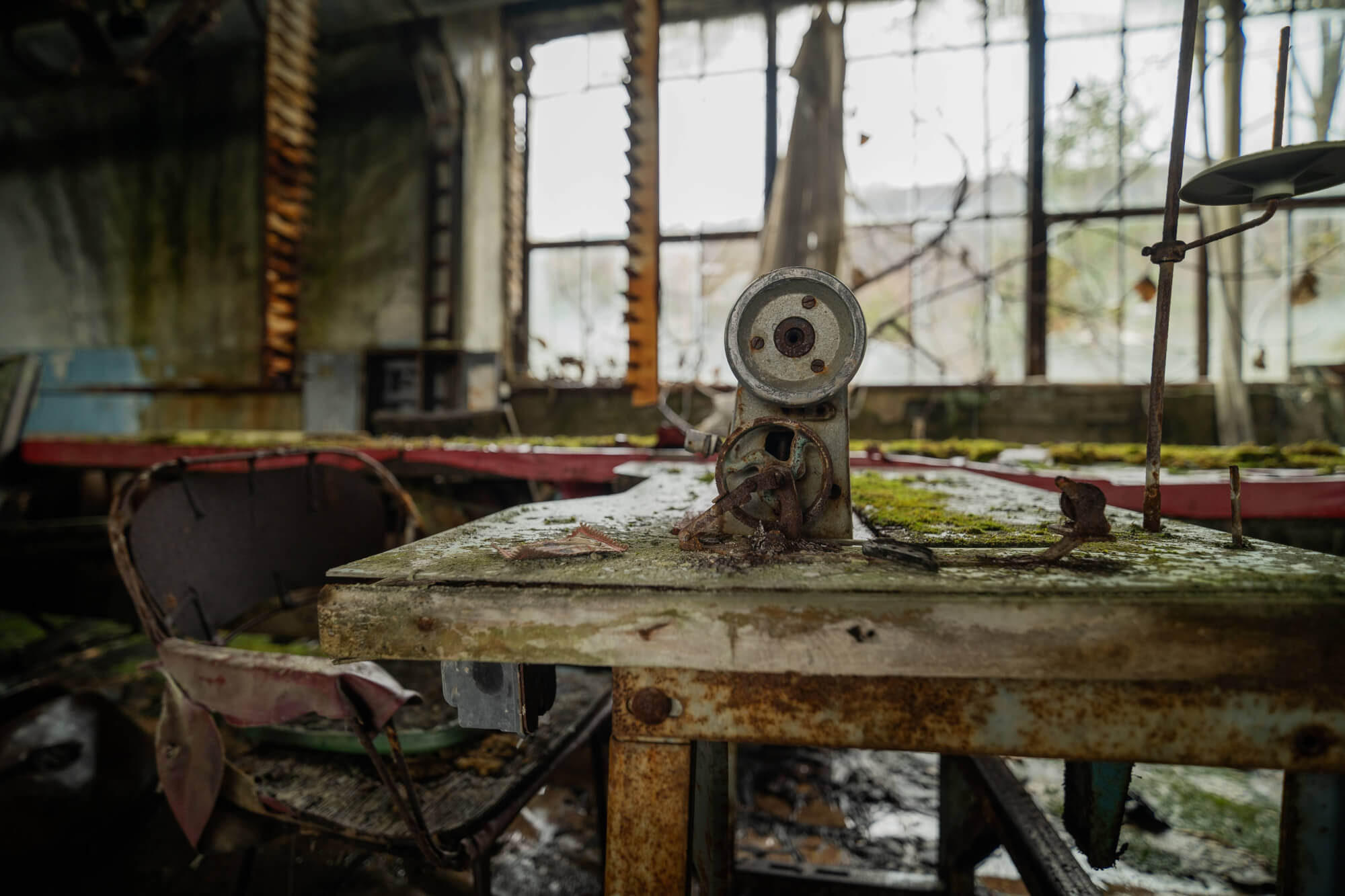 A close-up of a rusted sewing machine on a dilapidated table in an abandoned workshop, surrounded by debris and overgrown moss, with broken windows in the background.