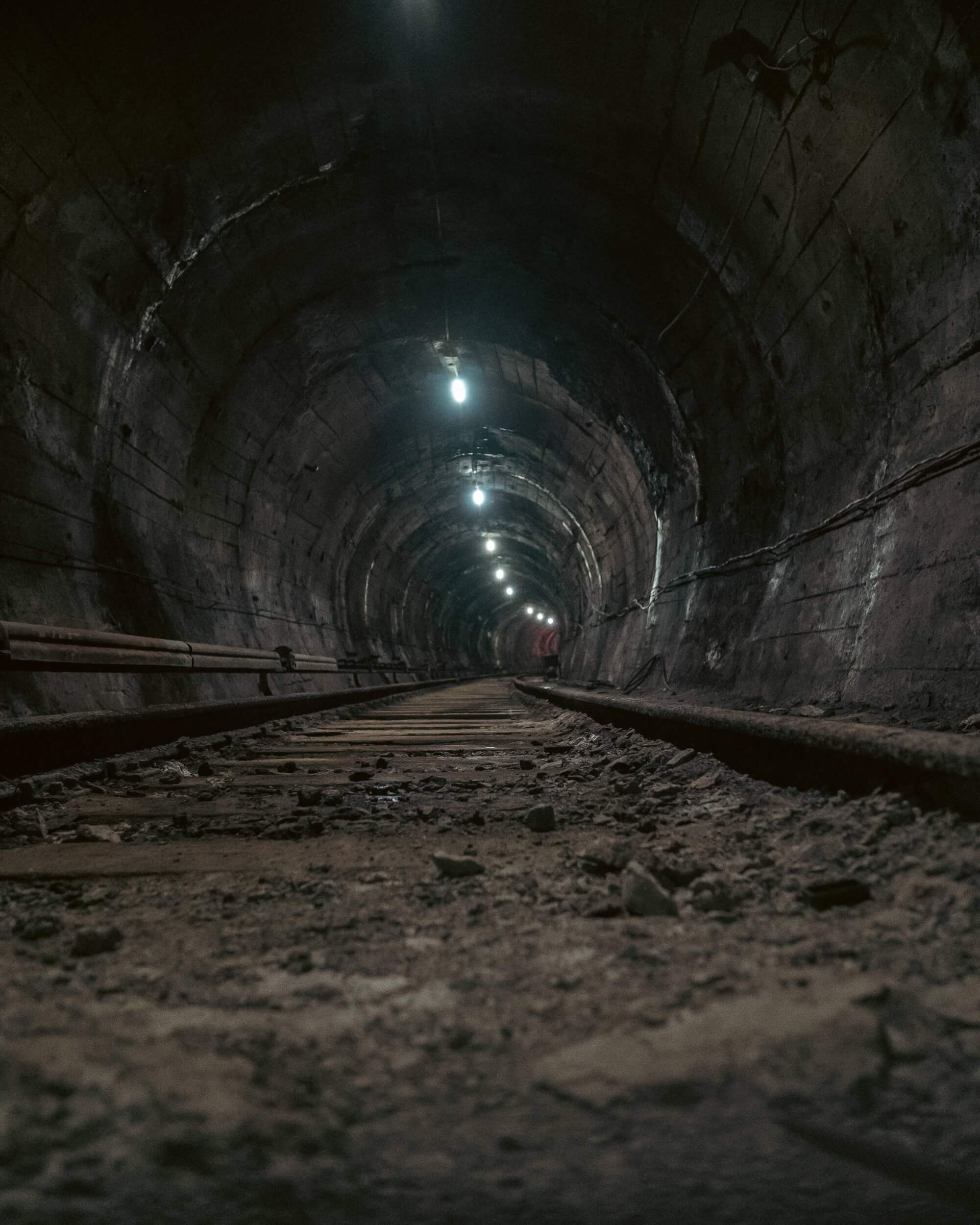 A perspective view of a long, dark tunnel with uneven walls and light bulbs illuminating the path. Wooden railway tracks are visible in the foreground covered with dust and debris.