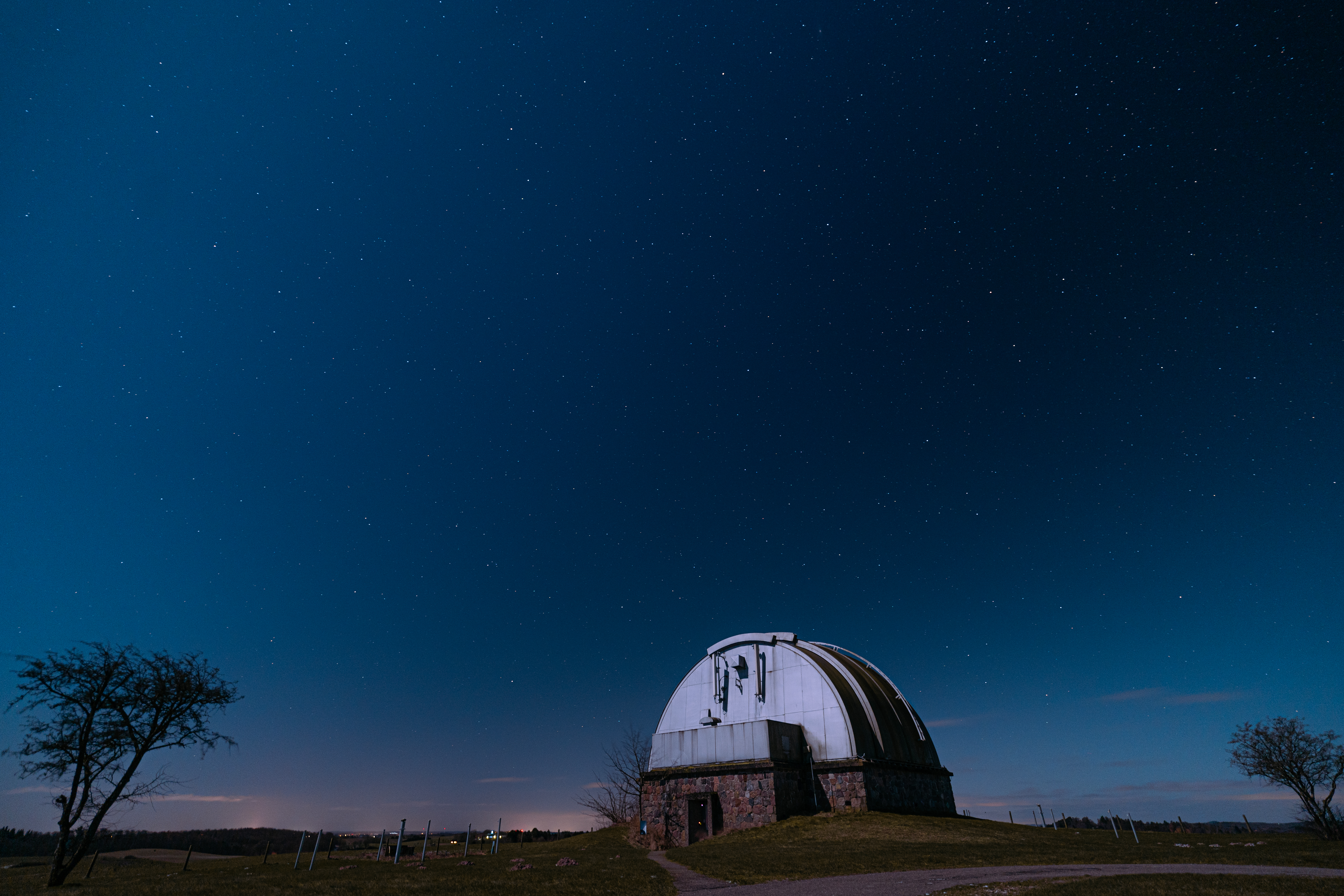 An observatory dome silhouetted against a starry night sky, with a faint glow on the horizon and sparse trees nearby.