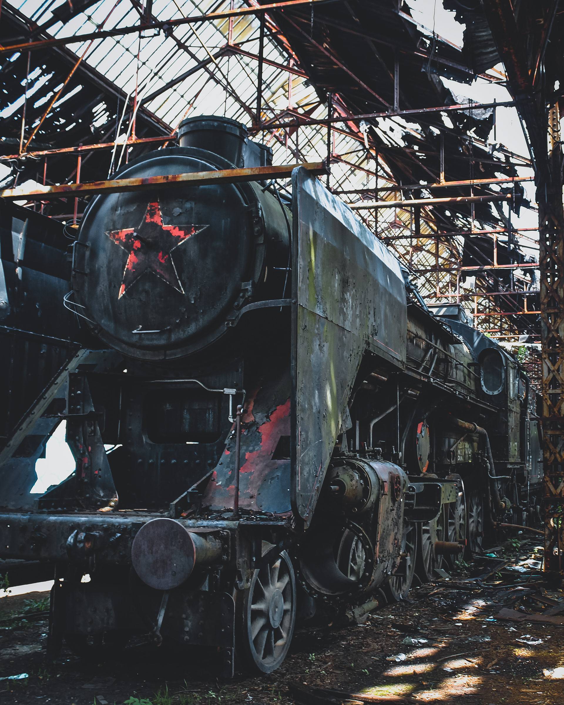 A weathered steam locomotive in an abandoned railway shed, with a red star emblem, surrounded by shadows and sunlight filtering through a broken roof.