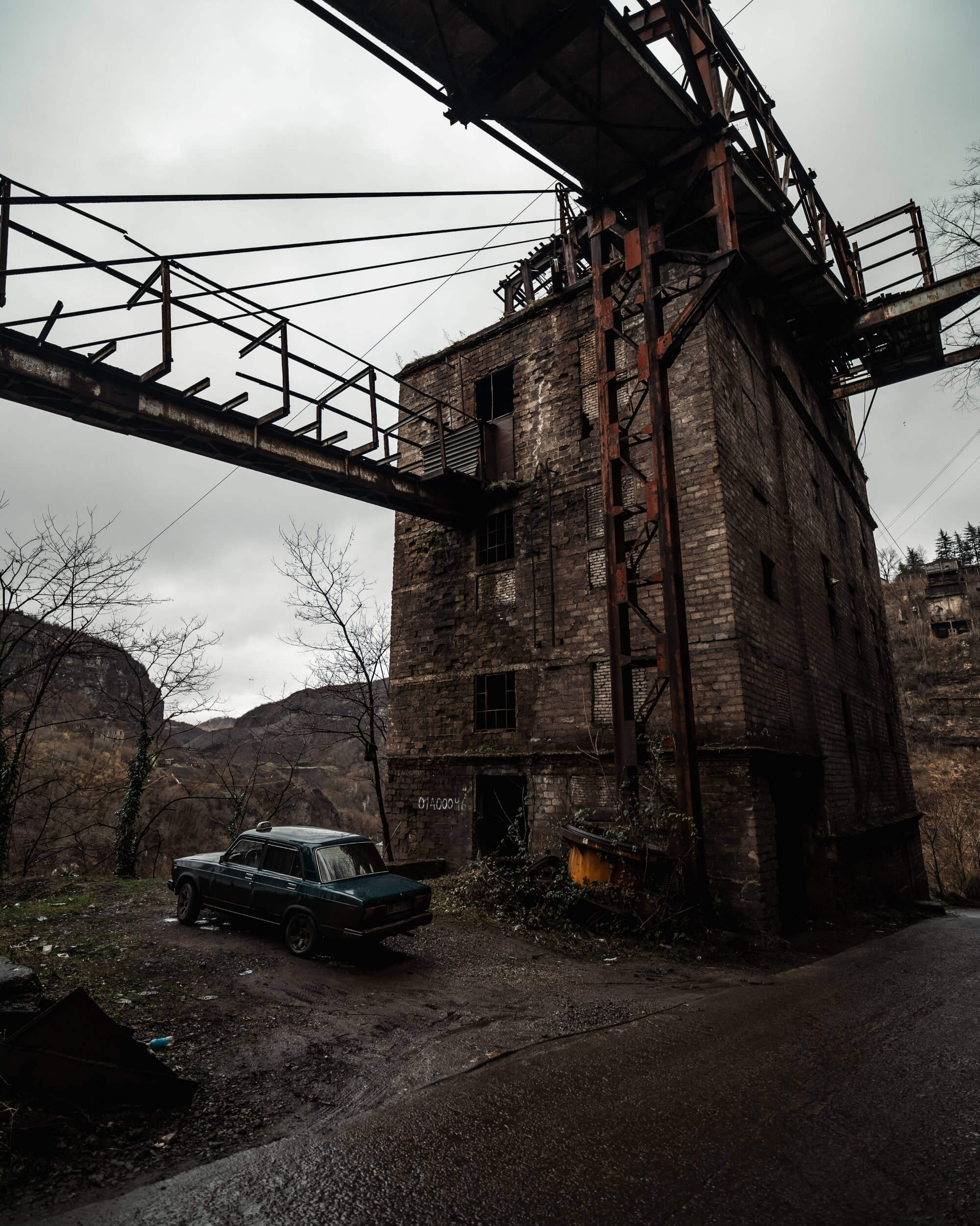 A weathered industrial building with a rusted walkway above it, alongside a parked vintage car on a muddy path, under a cloudy sky.