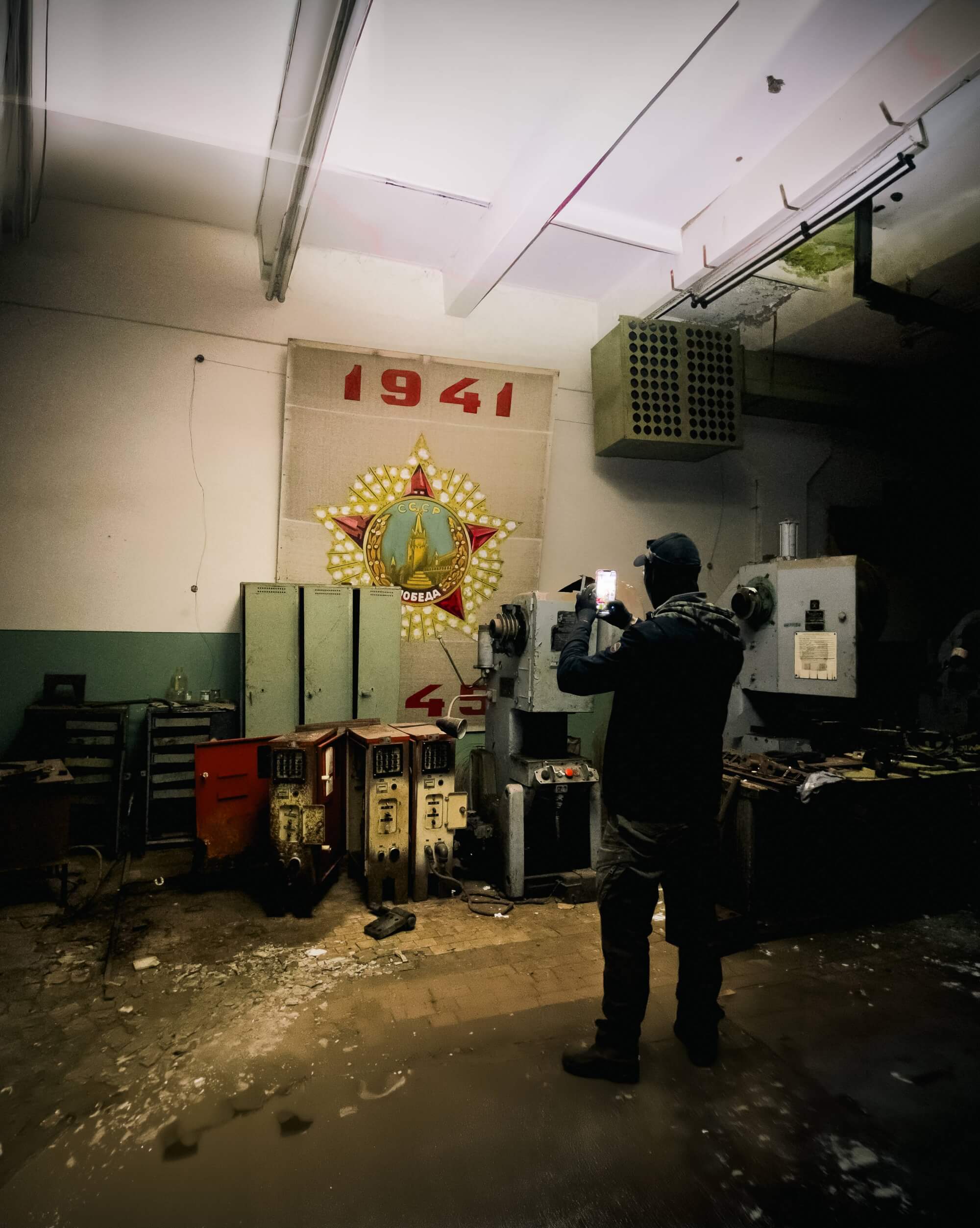 A person photographing a faded mural featuring a star and the year 1941 in an abandoned industrial setting, with scattered debris and old machinery around.