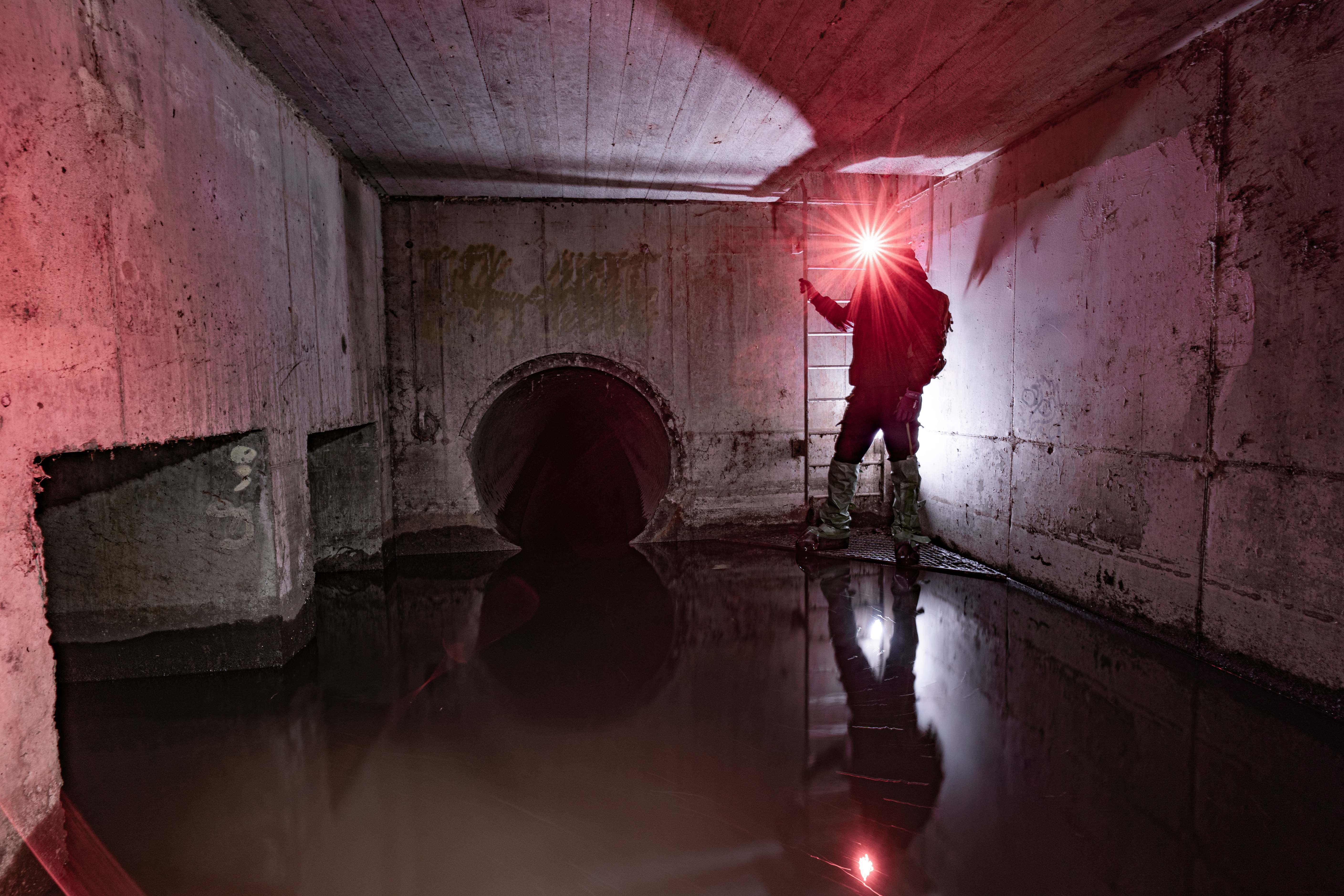 A person in dark clothing stands by a ladder in a dimly lit underground chamber with concrete walls. Water covers the floor, reflecting the light from a flashlight, creating an eerie atmosphere.
