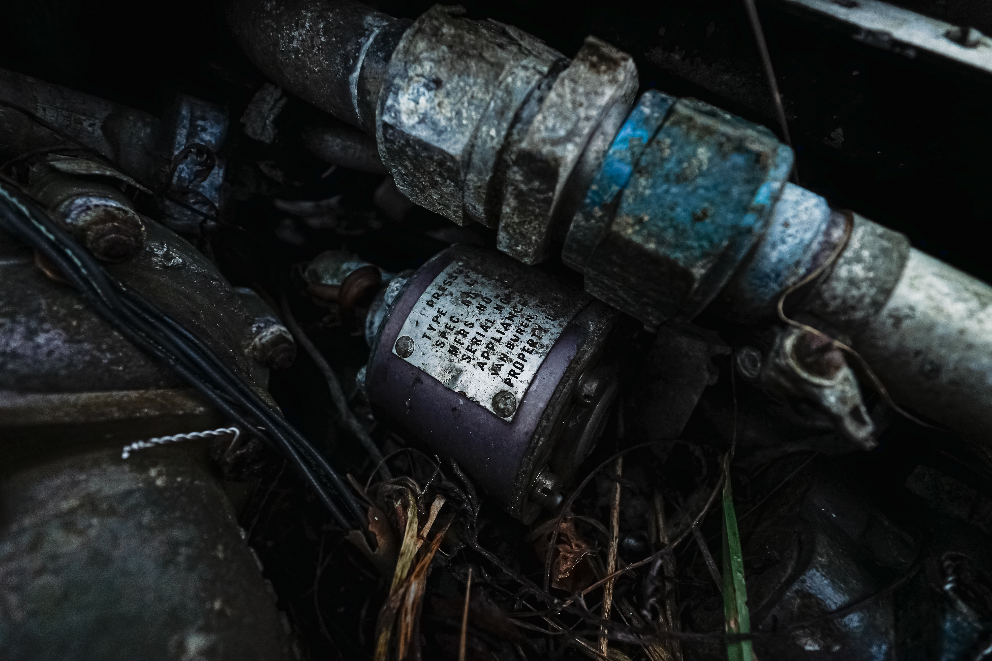 A close-up view of rusted mechanical parts with a focus on a purple cylindrical component and metal pipes. There is dry grass and debris surrounding the machinery, conveying a sense of neglect.