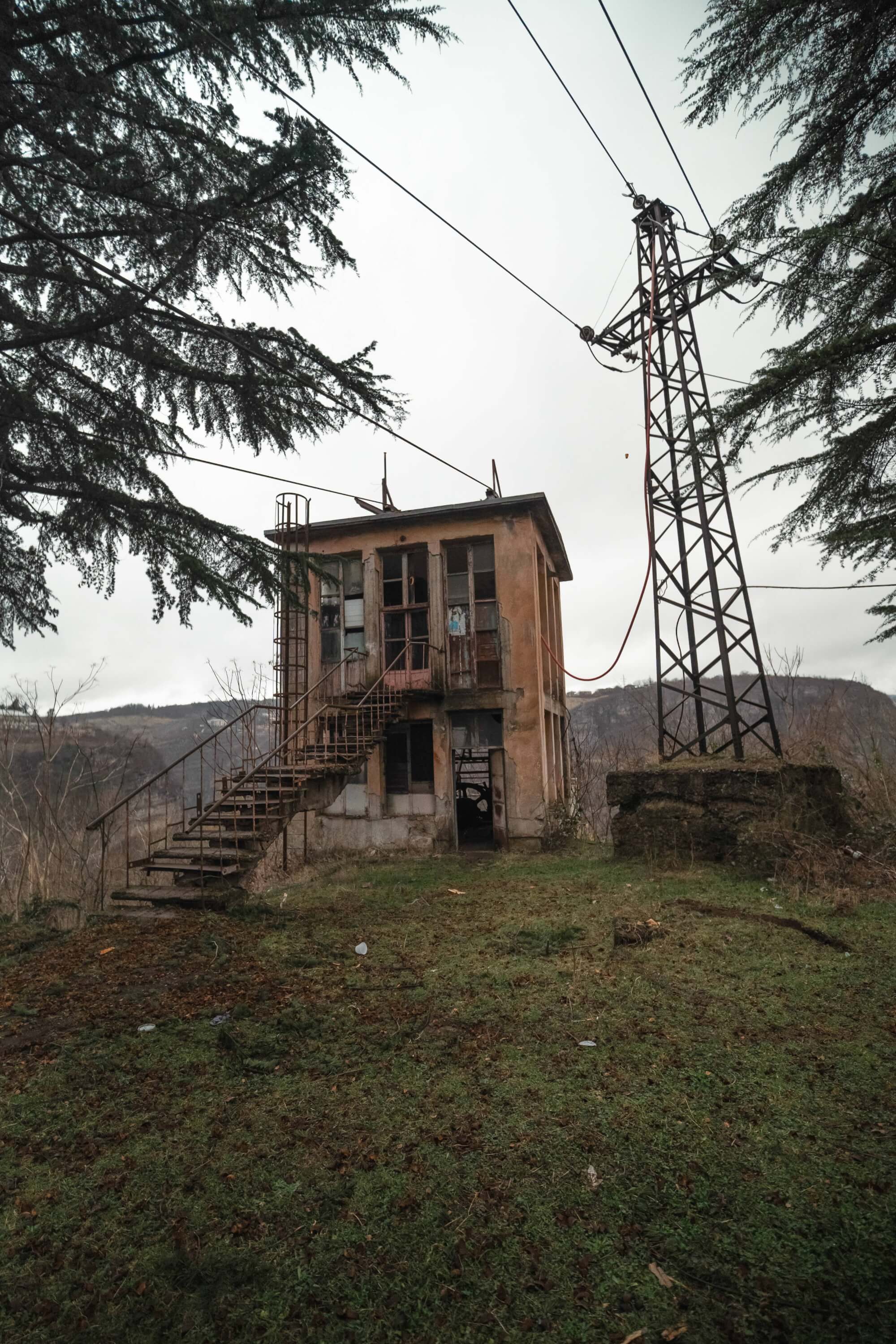 An abandoned building with peeling paint and broken windows, a staircase leading up to its entrance, next to a power pole. The scene is surrounded by sparse vegetation and dark trees under an overcast sky.