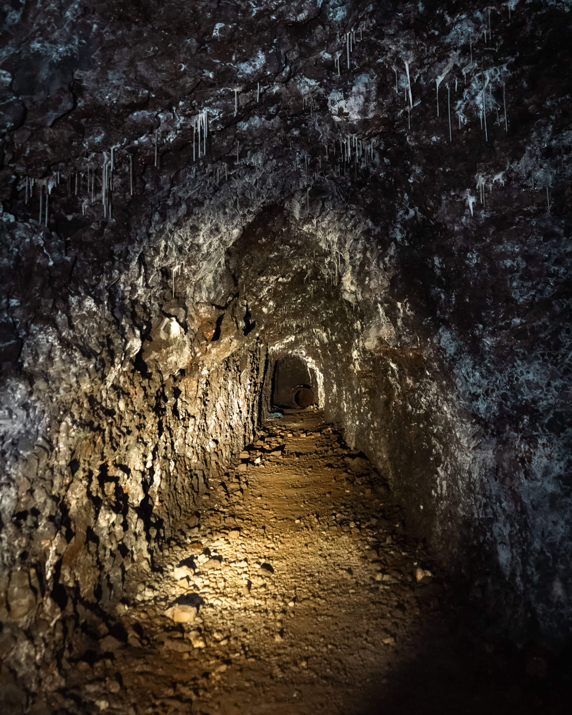 A narrow tunnel in a cave with rough, rocky walls and visible stalactites. The ground is uneven with scattered rocks and dirt, illuminated by a soft light coming from the side.