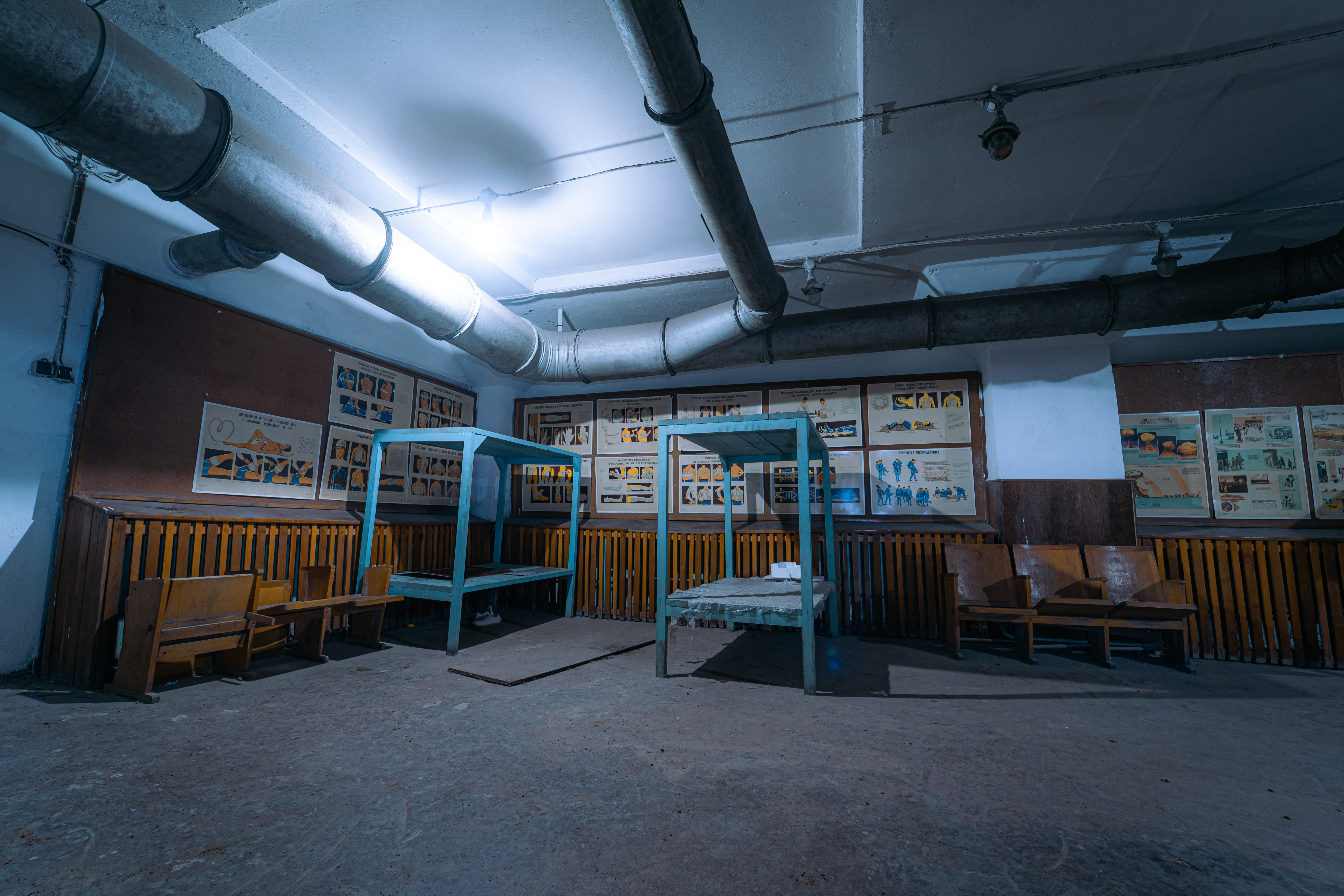 An abandoned room featuring two empty wooden beds and educational posters on the walls. The space is illuminated by cool blue lighting, and overhead metal pipes are visible.