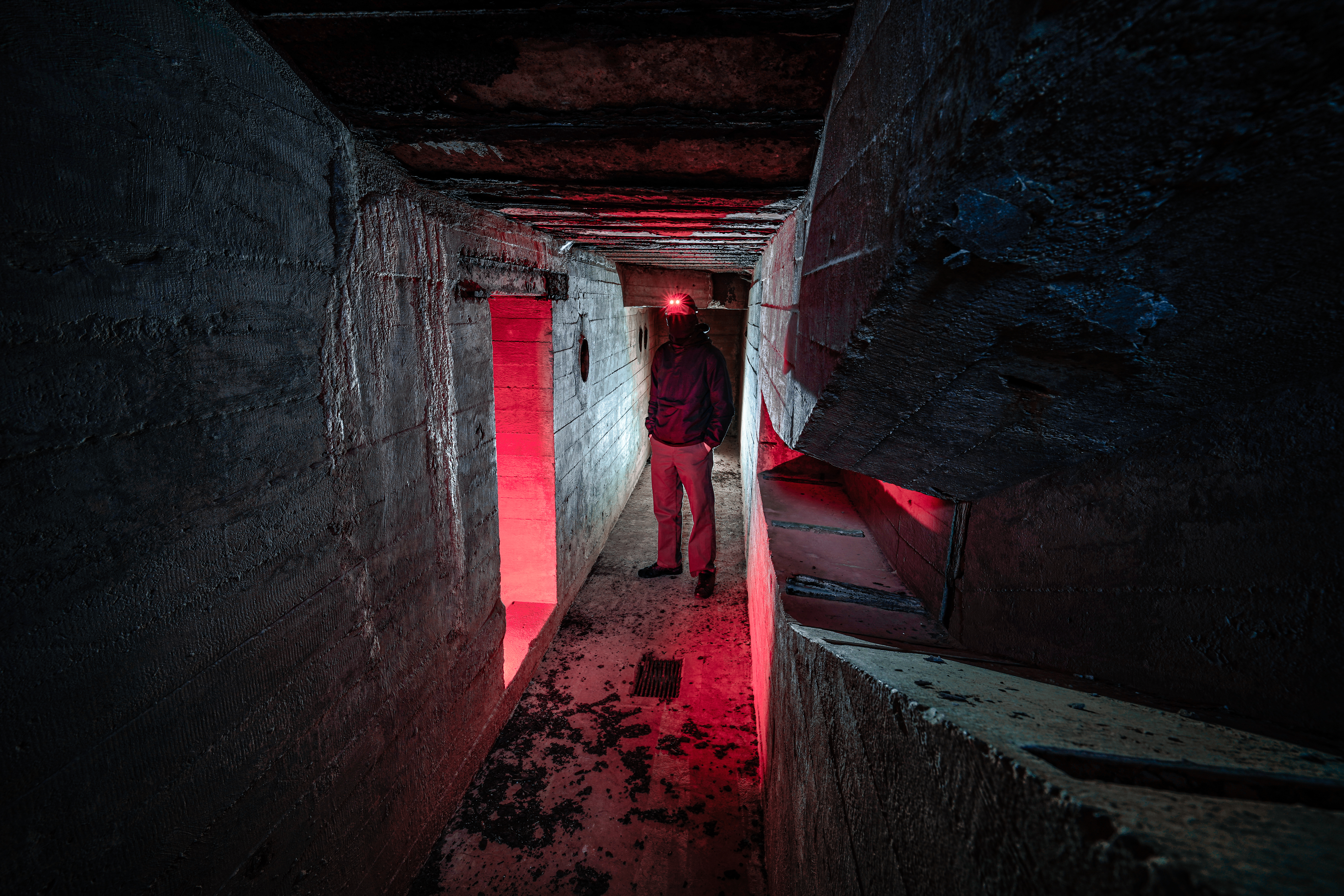 A person standing in a dark, narrow concrete corridor with red lighting illuminating an entrance ahead.