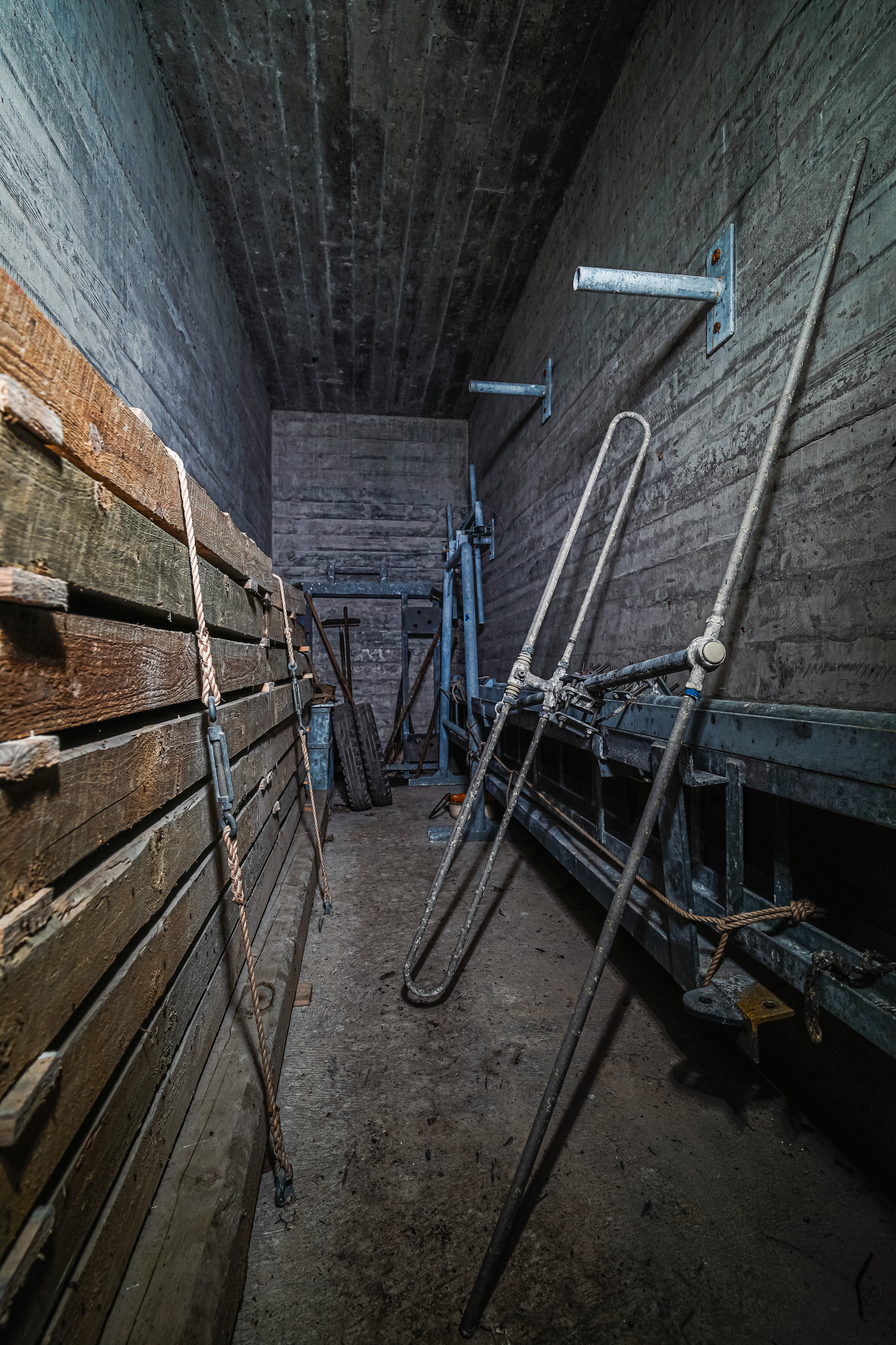 A dimly lit, narrow room with wooden beams stacked against the left wall and various metal structures on the right, including a large frame and ropes. The concrete walls and floor add to the industrial feel of the space.
