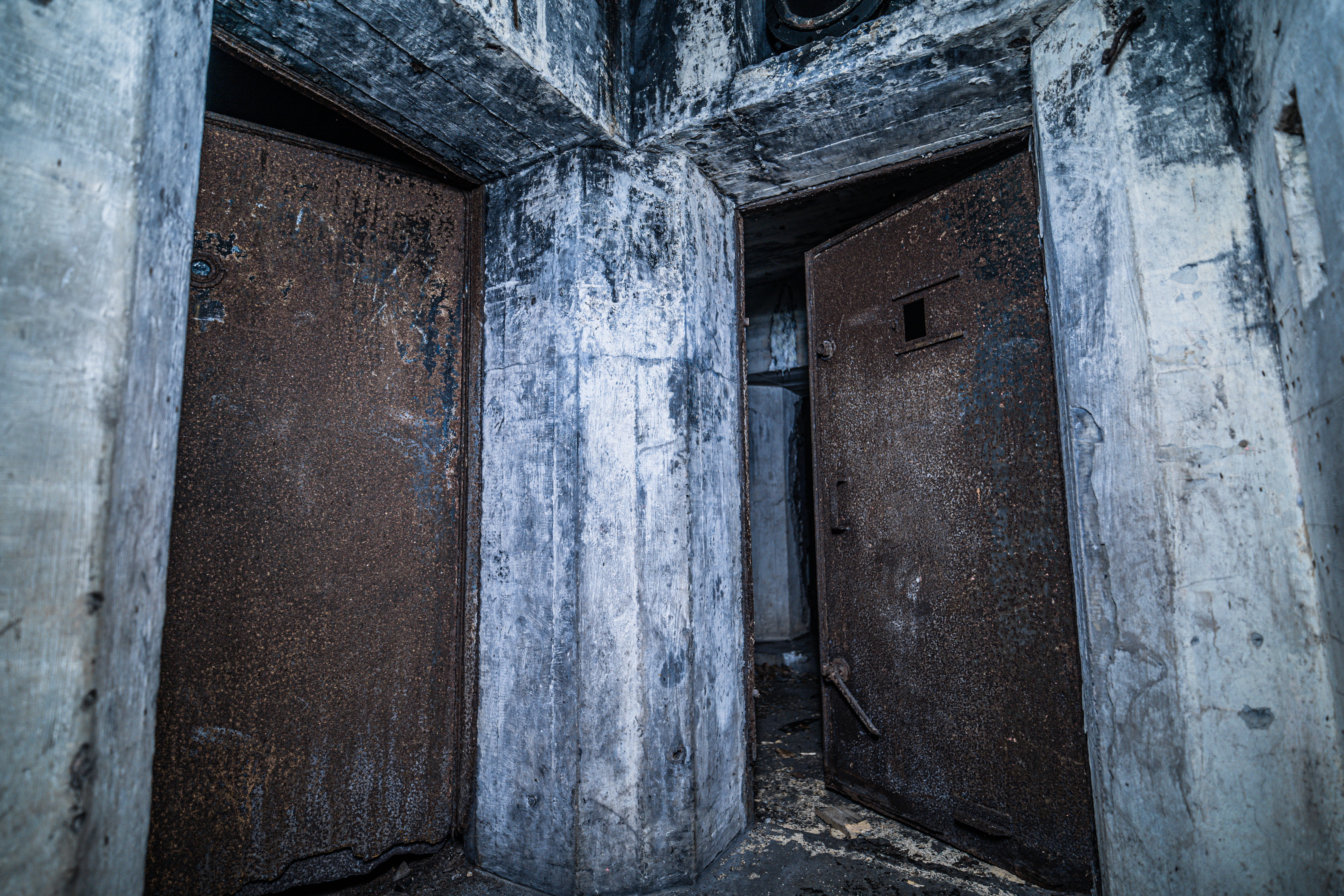 An interior view featuring two rusted metal doors on either side of a concrete column, with weathered concrete walls. The scene is dimly lit, creating dramatic shadows.