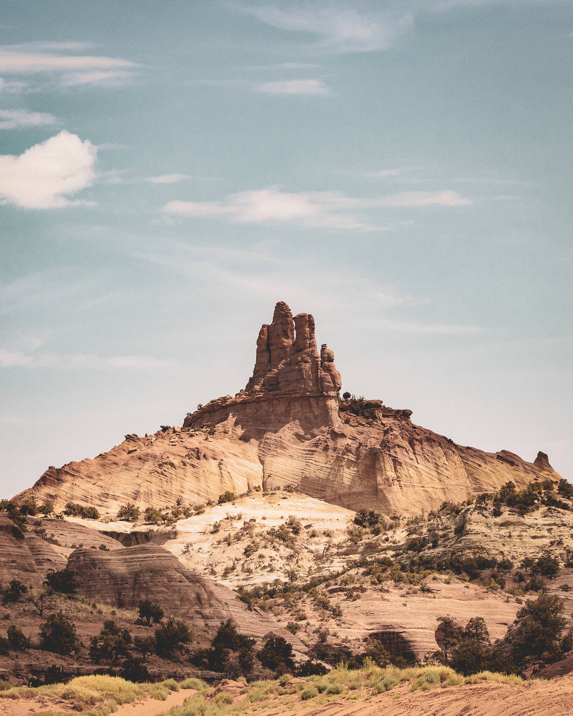 A towering rock formation rises majestically against a blue sky with wispy clouds, surrounded by rolling hills and patches of green vegetation.