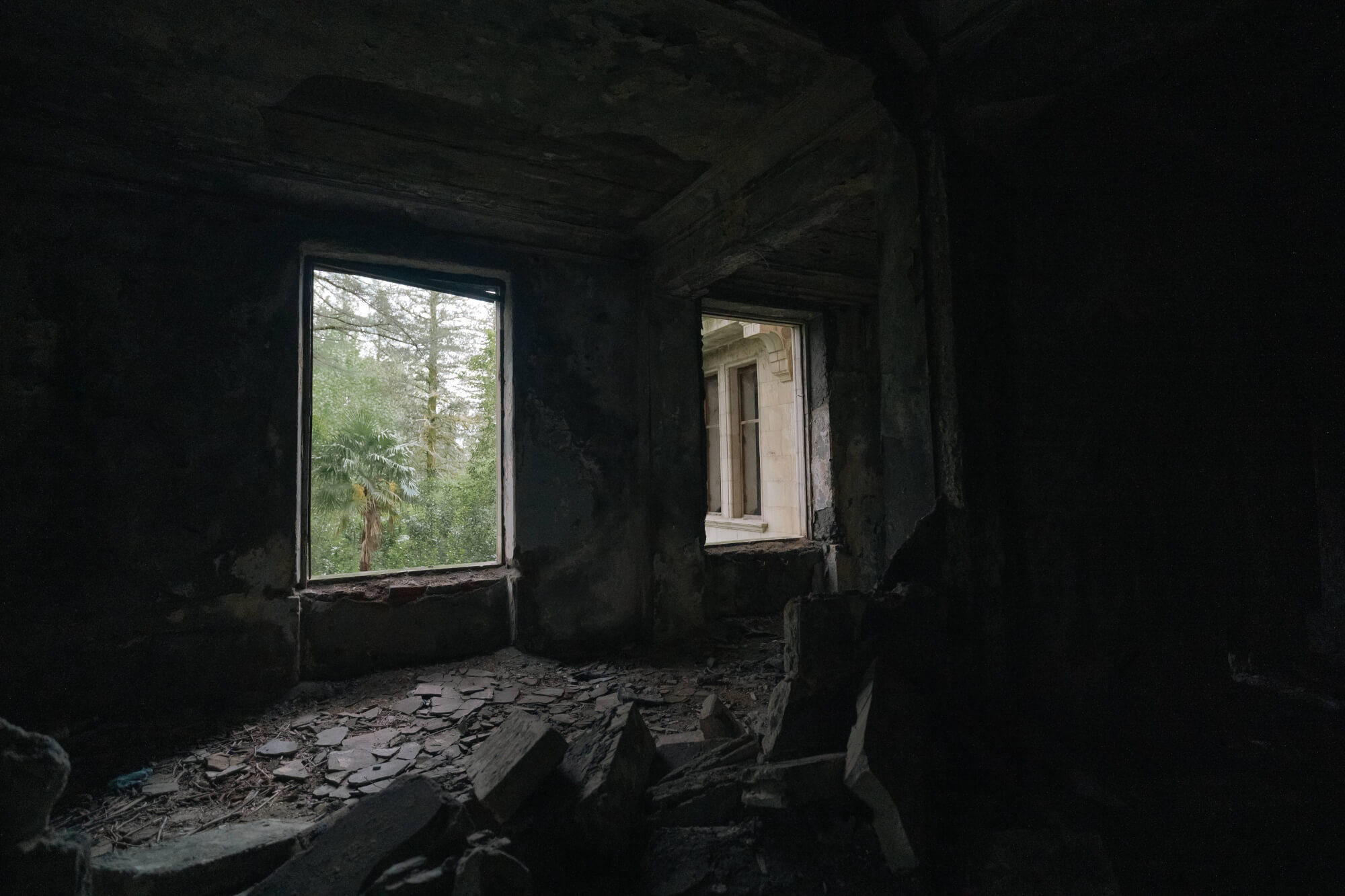 Interior of an abandoned room with two windows. One window shows green trees outside, while the other reveals an exterior wall. The room has a dark, dusty floor littered with stones and debris, with peeling walls.